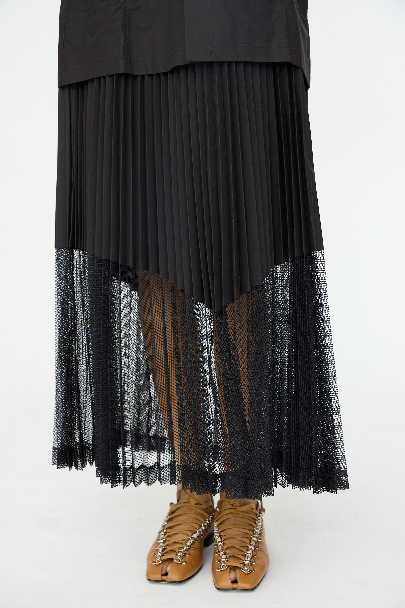 A person wears the TOGA ARCHIVES Typewriter Mesh Dress in Black, featuring a long, pleated silhouette with sheer lower section, paired with tan split-toe lace-up shoes, standing against a white background.