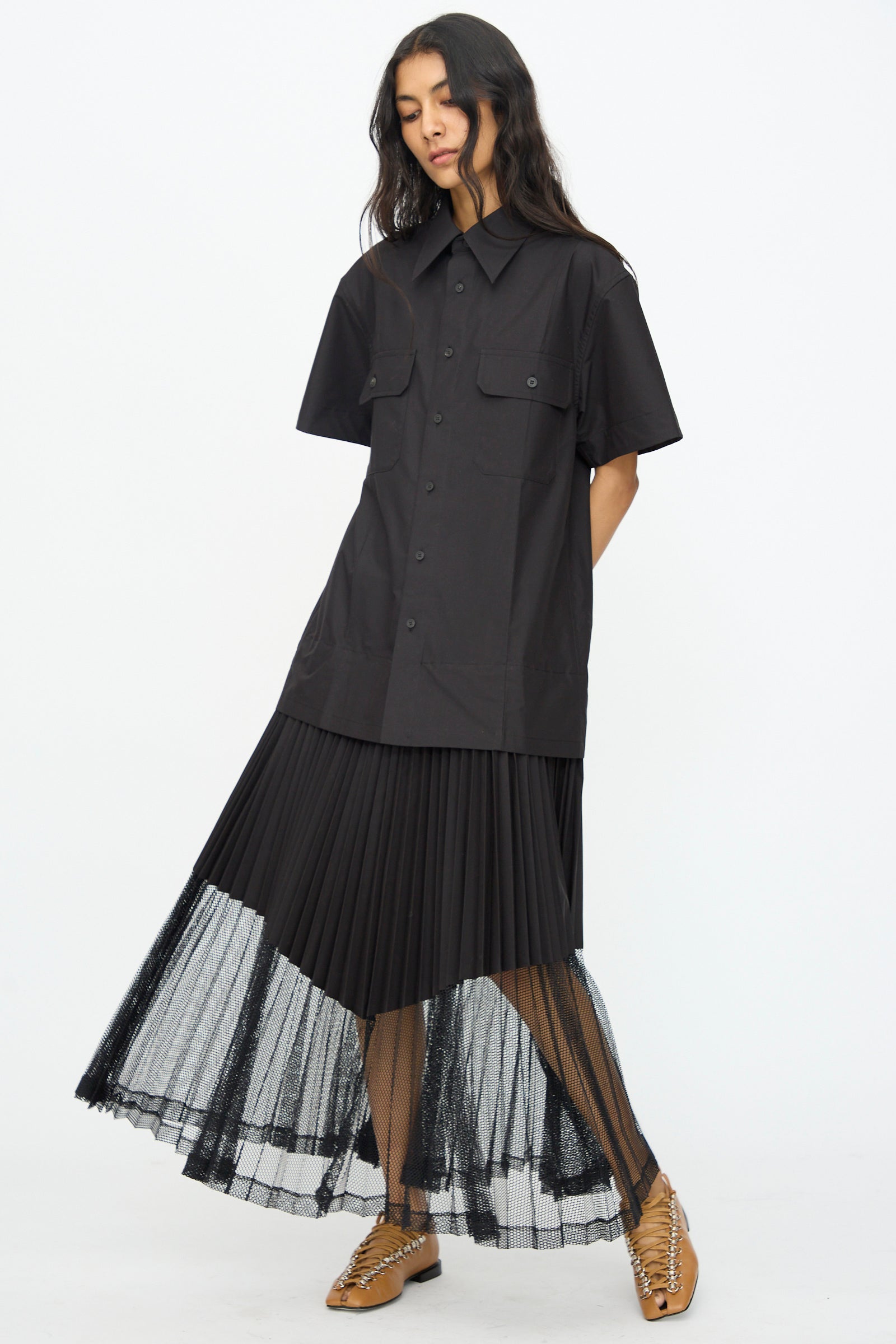 A woman wears the TOGA ARCHIVES Typewriter Mesh Dress in Black, featuring a short-sleeve button-up design with chest pockets and mesh panels, paired with tan lace-up shoes, standing against a white background.