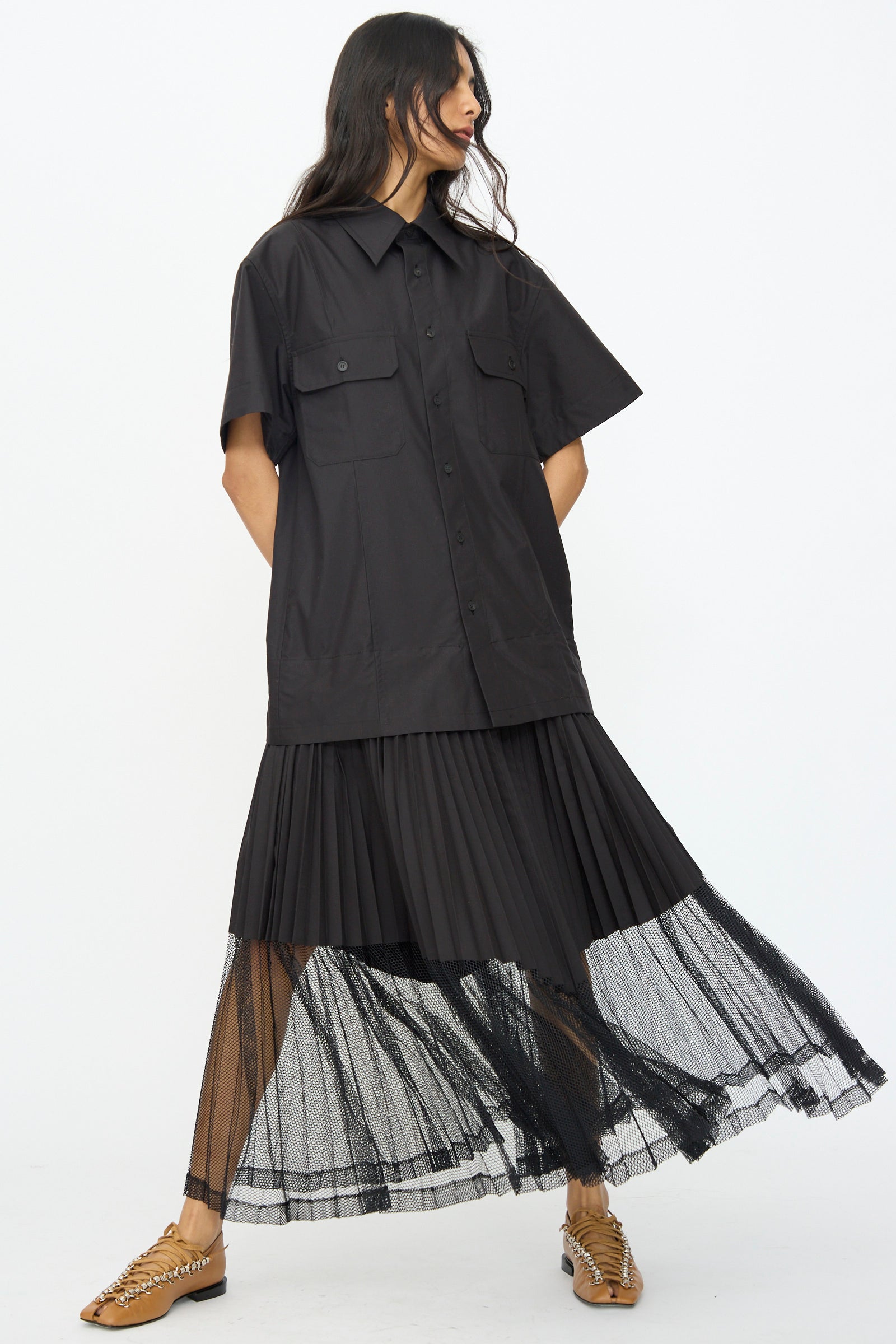 A person wearing the TOGA ARCHIVES Typewriter Mesh Dress in Black, featuring a short-sleeve button-up style with chest pockets and a mesh-hem pleated skirt, stands against a plain white background.