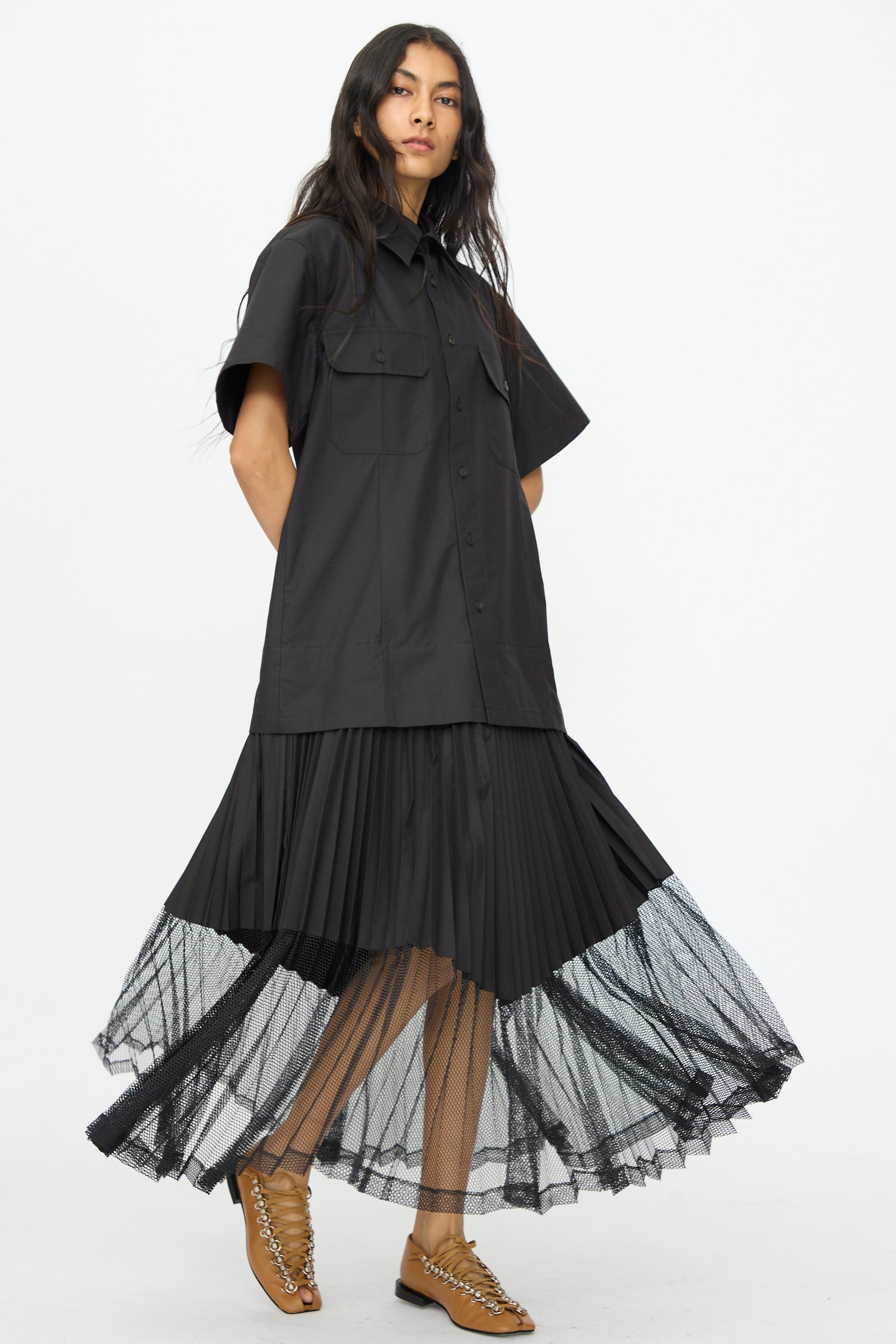 A woman wears the TOGA ARCHIVES Typewriter Mesh Dress in Black, featuring a pleated skirt with a sheer mesh hem, styled with tan open-toe lace-up shoes.