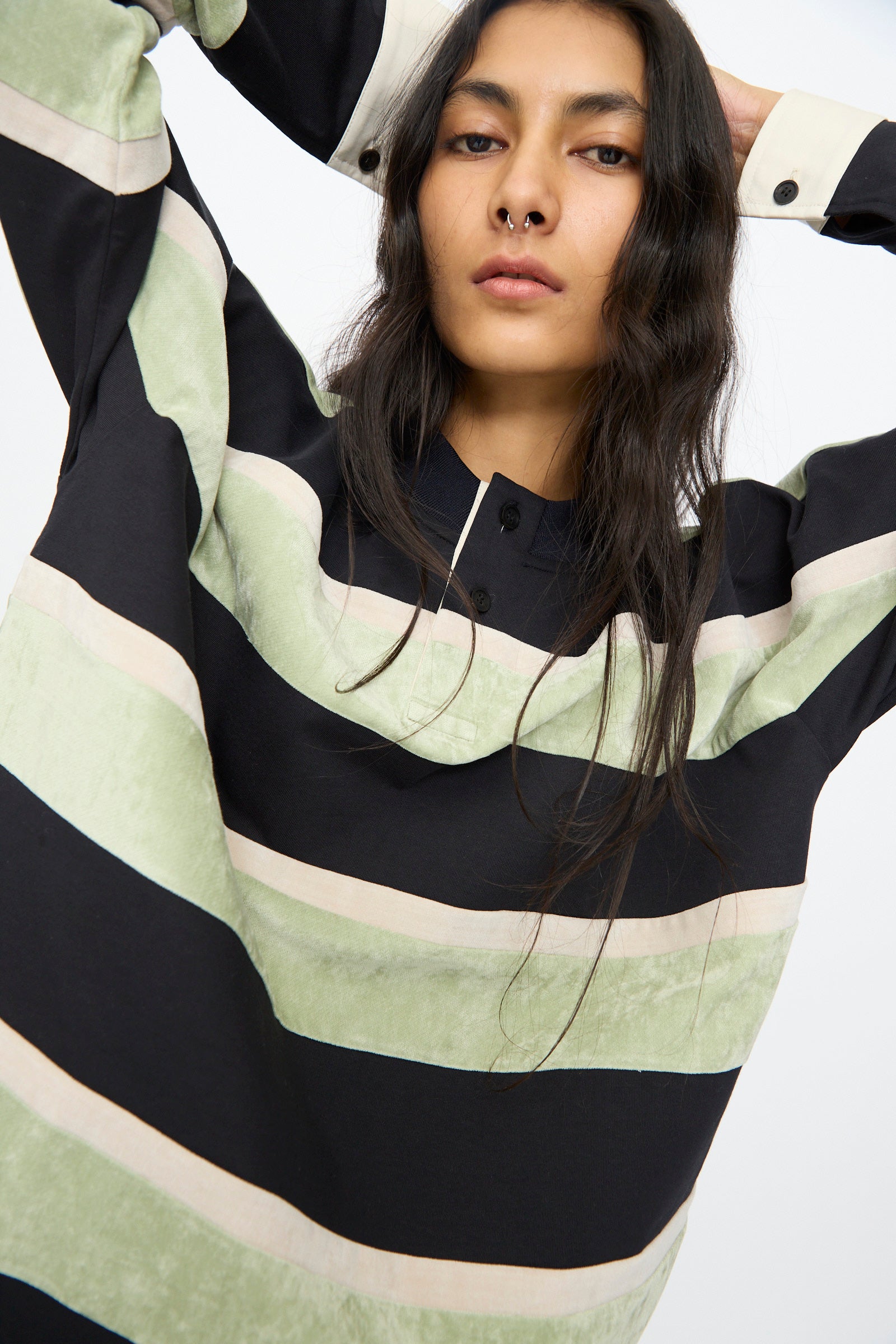 A woman with long dark hair wears the TOGA ARCHIVES Velvet Border Jersey Long Sleeve in Light Green, standing against a plain background with her arms raised behind her head.