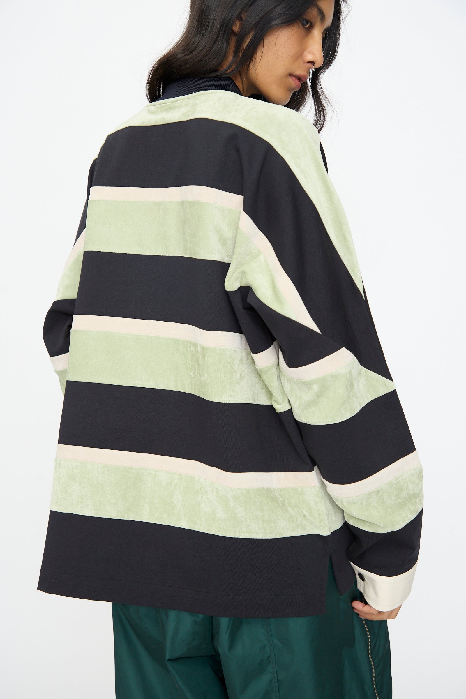 Person wearing the TOGA ARCHIVES Velvet Border Jersey Long Sleeve in Light Green, featuring wide black, light green, and beige stripes, viewed from the back. The person has long dark hair and green pants.