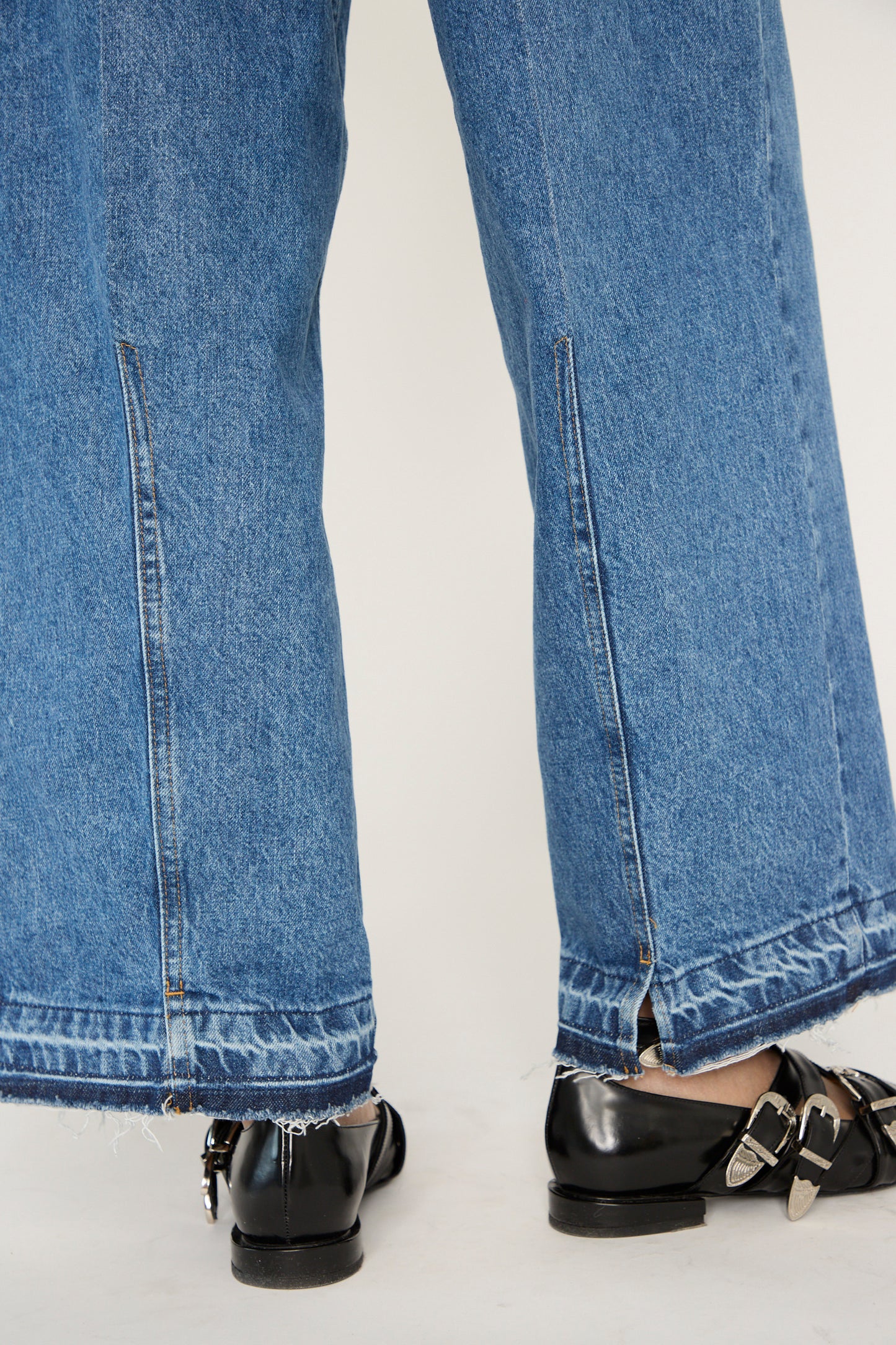 Close-up of the lower back of a person wearing TOGA ARCHIVES Wide Denim Pant in Blue with black shoes featuring silver buckles.