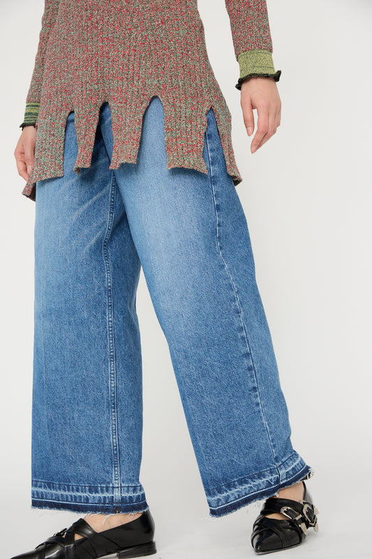 Person wearing a knit top with a jagged hem, Wide Denim Pant in Blue by TOGA ARCHIVES, and black pointed shoes against a white background. 