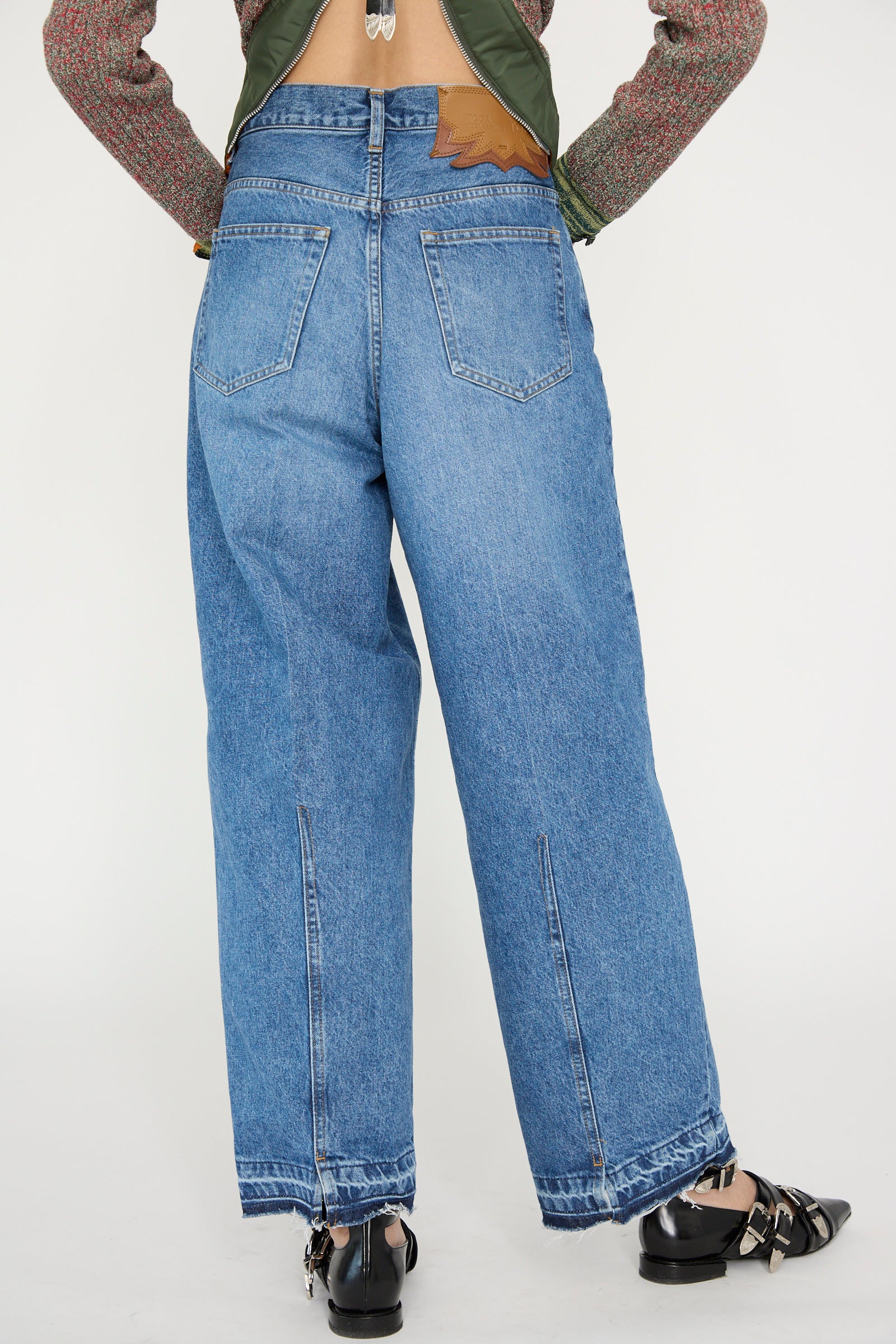 A person in TOGA ARCHIVES Wide Denim Pant in Blue and black shoes stands with their back to the camera, hands on hips against a plain backdrop. 