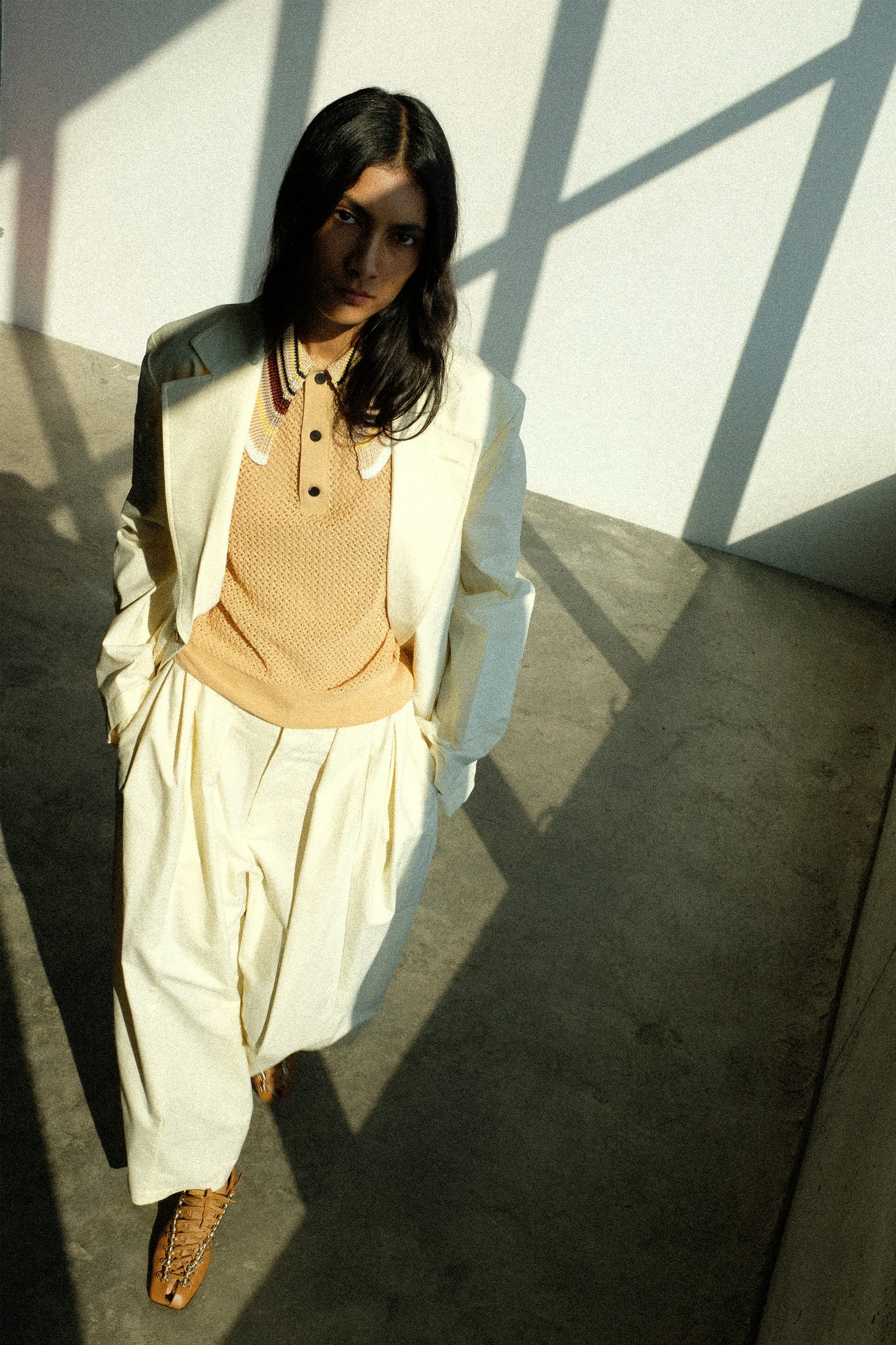 Woman in Toga Archives Ox Suit with the Lace Knit Polo Shirt in Beige, walking against a white wall with shadows.