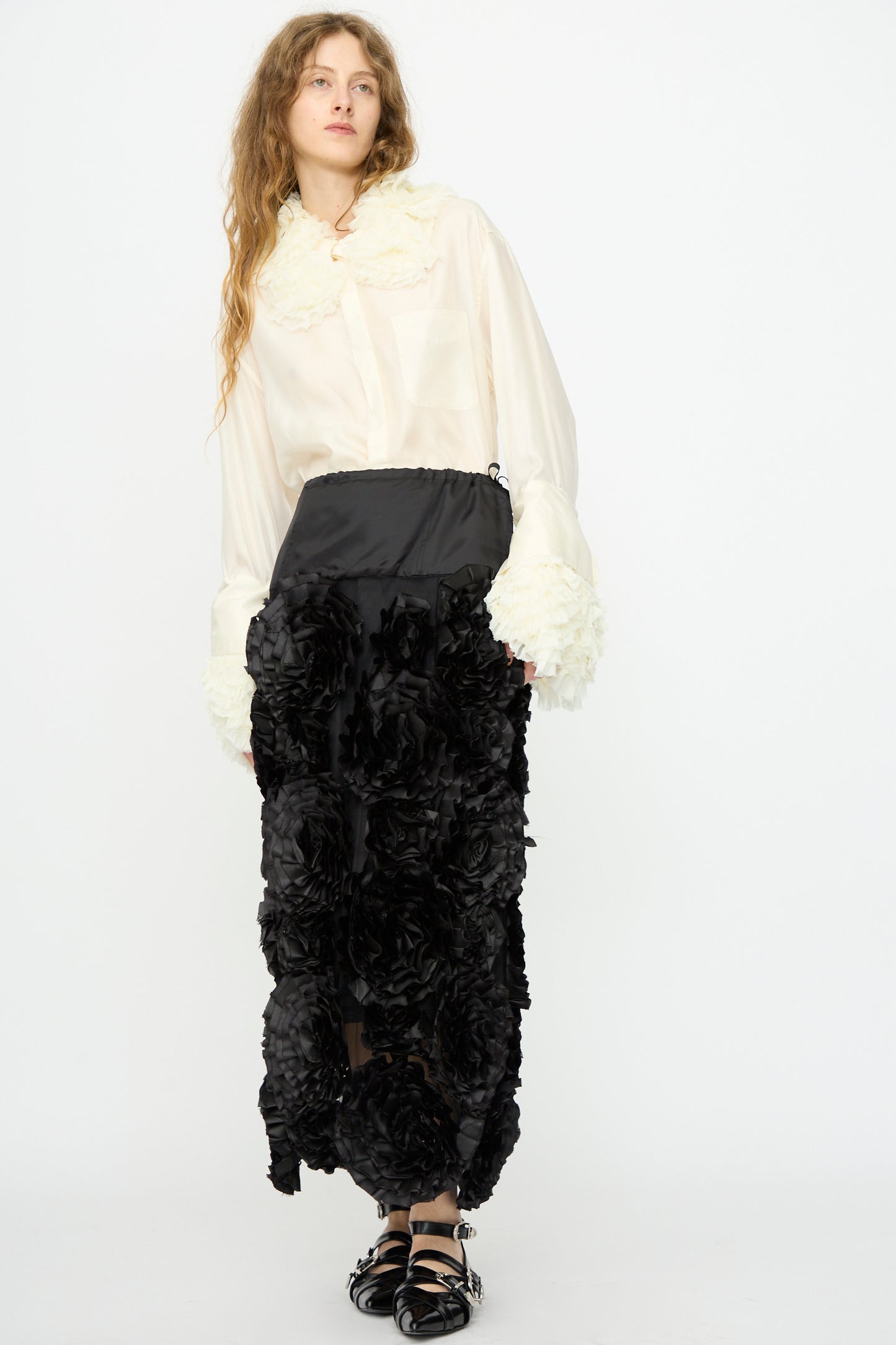 A woman stands against a plain white background wearing a white blouse and the Frill Embroidery Skirt in Black by TOGA ARCHIVES, styled with black sandals.