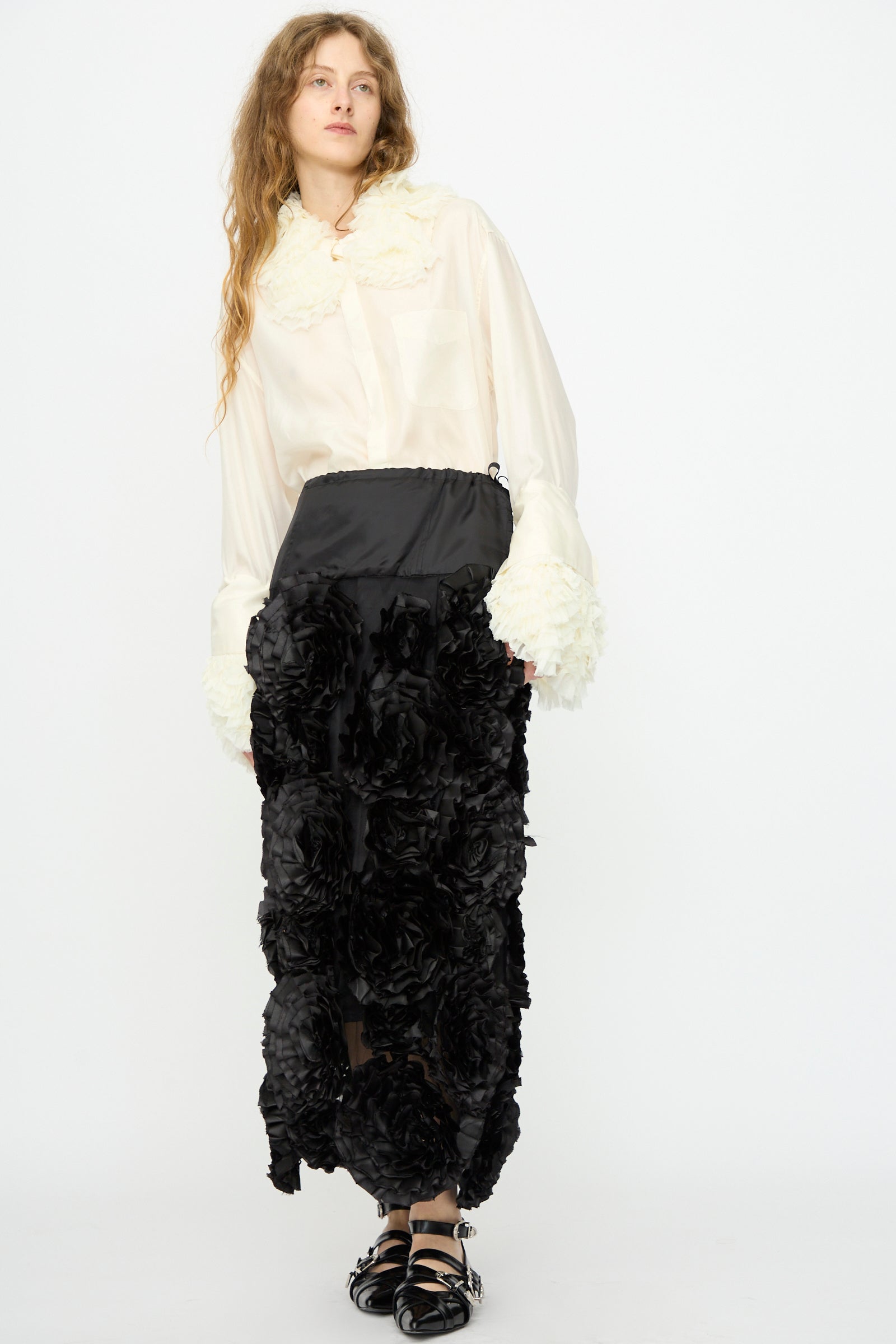 A woman stands against a plain white background wearing a white blouse and the Frill Embroidery Skirt in Black by TOGA ARCHIVES, styled with black sandals.