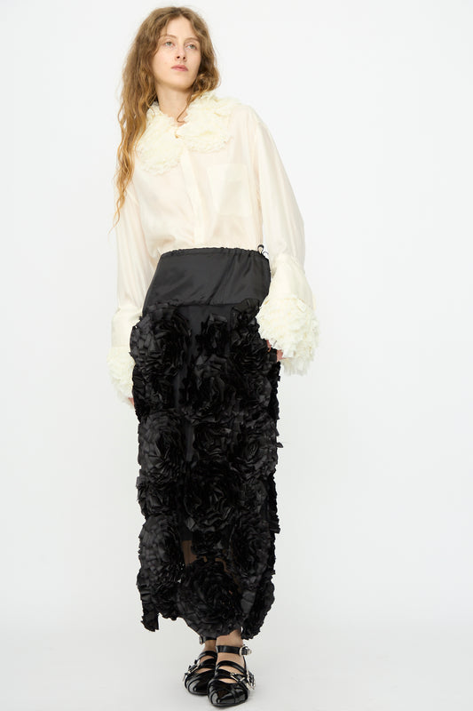 A woman stands against a plain white background wearing a white blouse and the Frill Embroidery Skirt in Black by TOGA ARCHIVES, styled with black sandals.