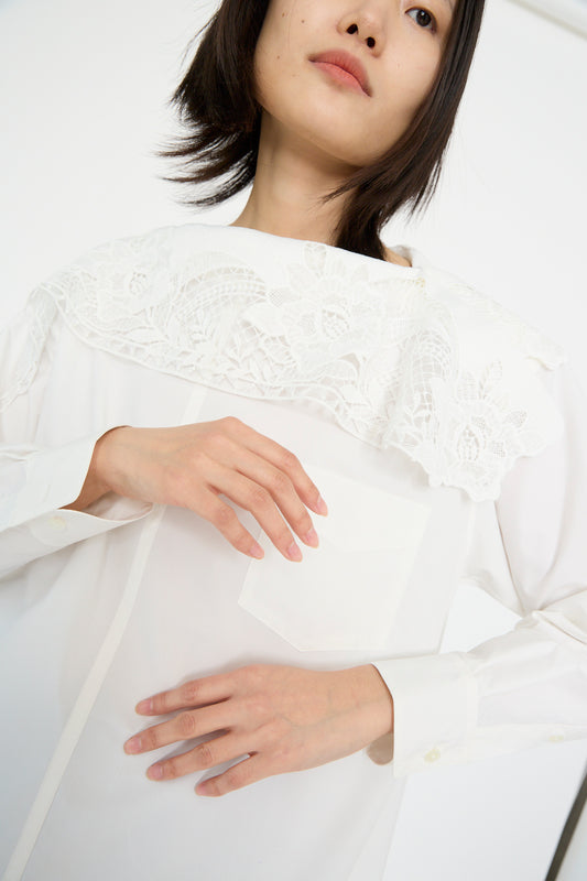 A person is wearing a Lace Boat Neck Shirt in White by TOGA ARCHIVES, with lace detail on the shoulders, holding their hands near their chest.