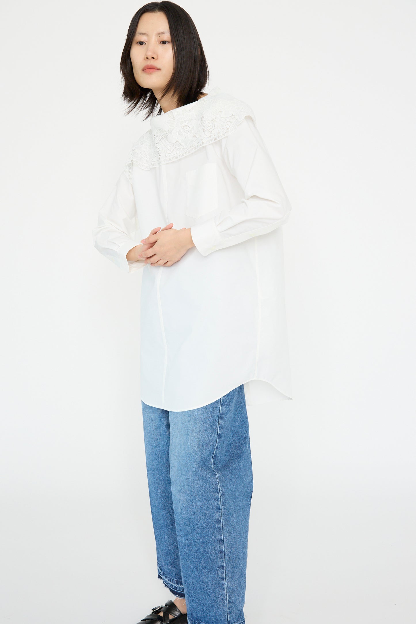 An individual showcases a Lace Boat Neck Shirt in White by TOGA ARCHIVES and blue wide-leg jeans against a plain white background.