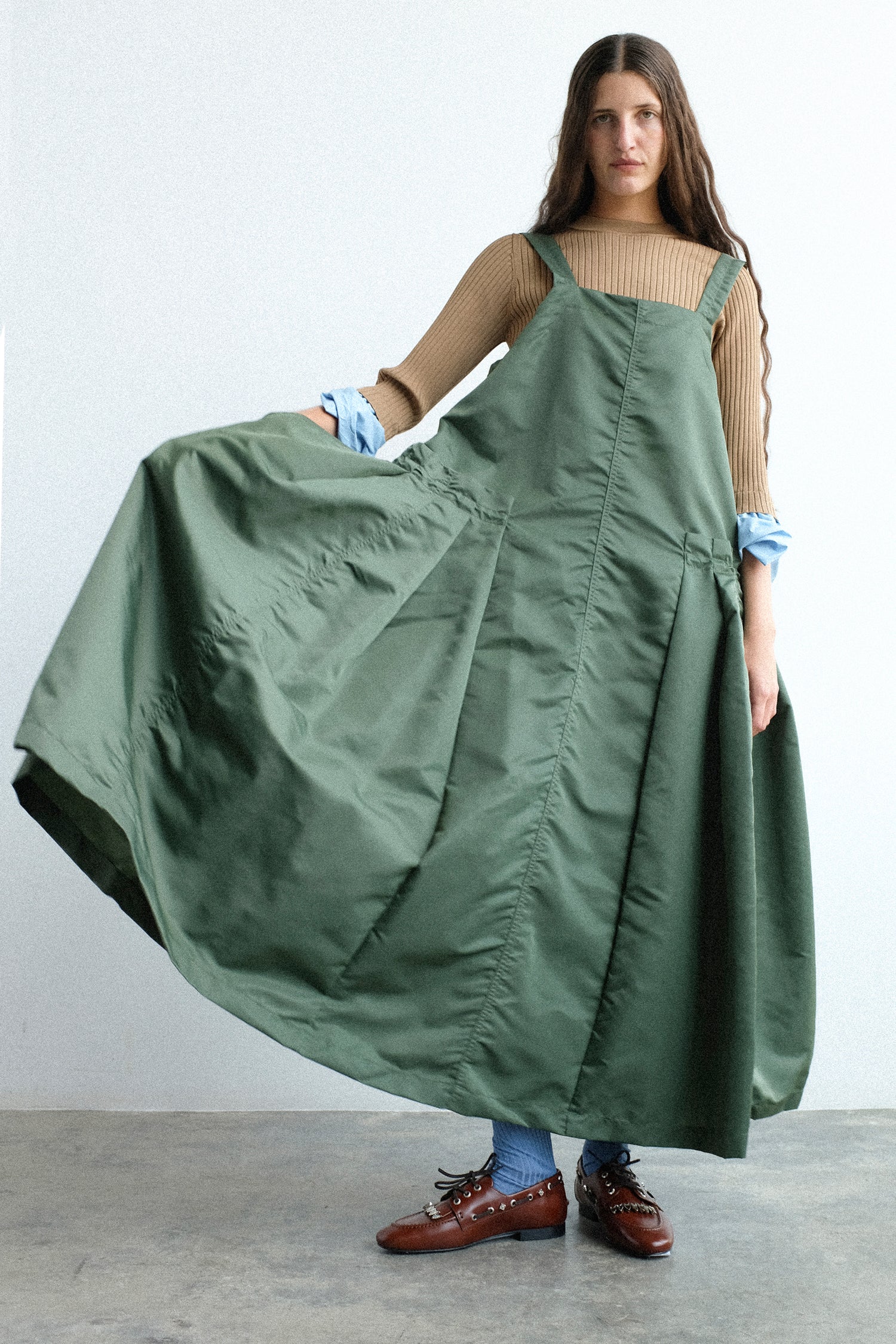 A model wearing Toga Archives Nylon Twill Dress in Khaki with blue socks and Leather Flat in Brown, holding the dress up, against a white background.