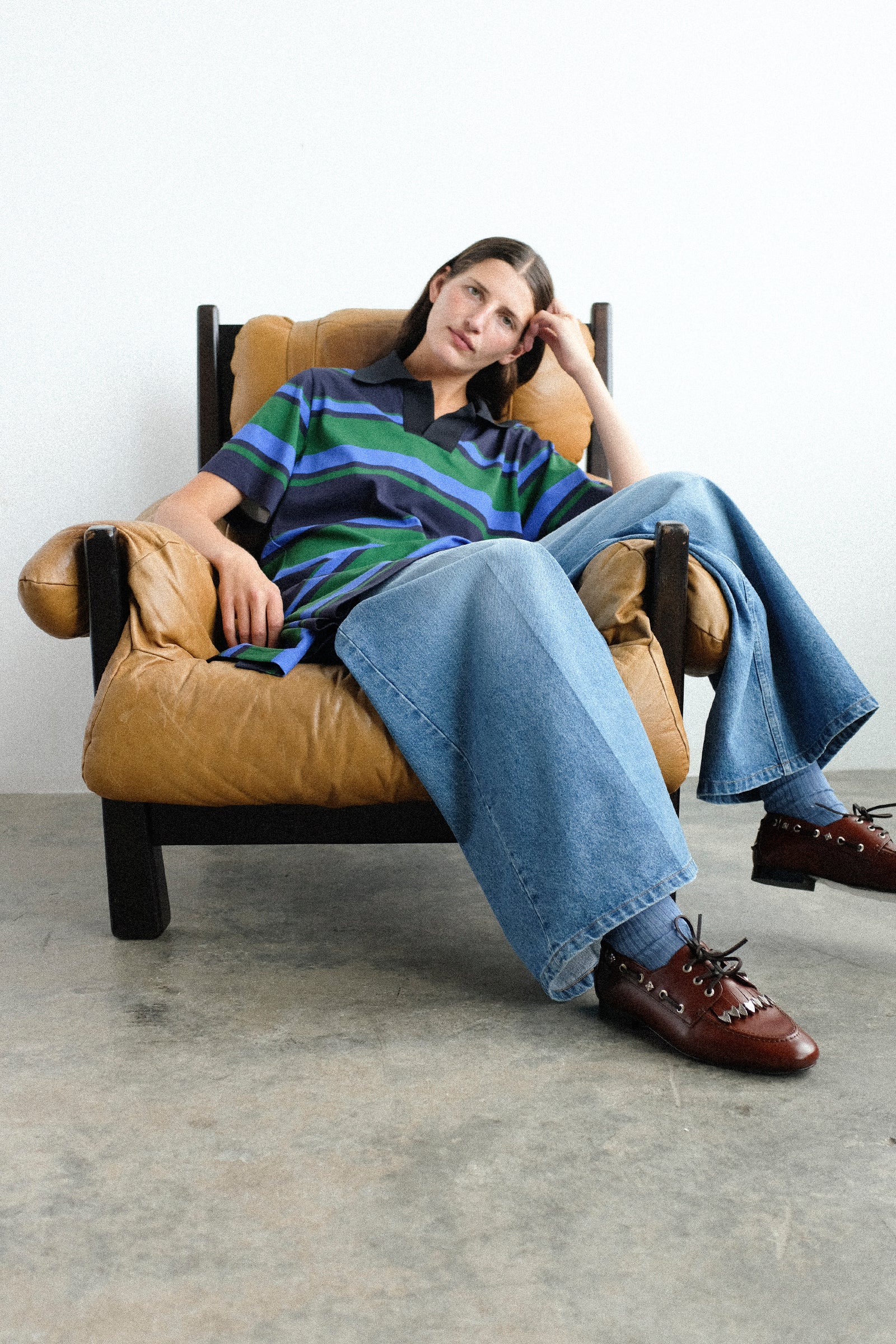 A person sitting in a tan leather armchair wearing Toga Archives Border Print Dress in Navy, against a plain white wall and concrete floor.
