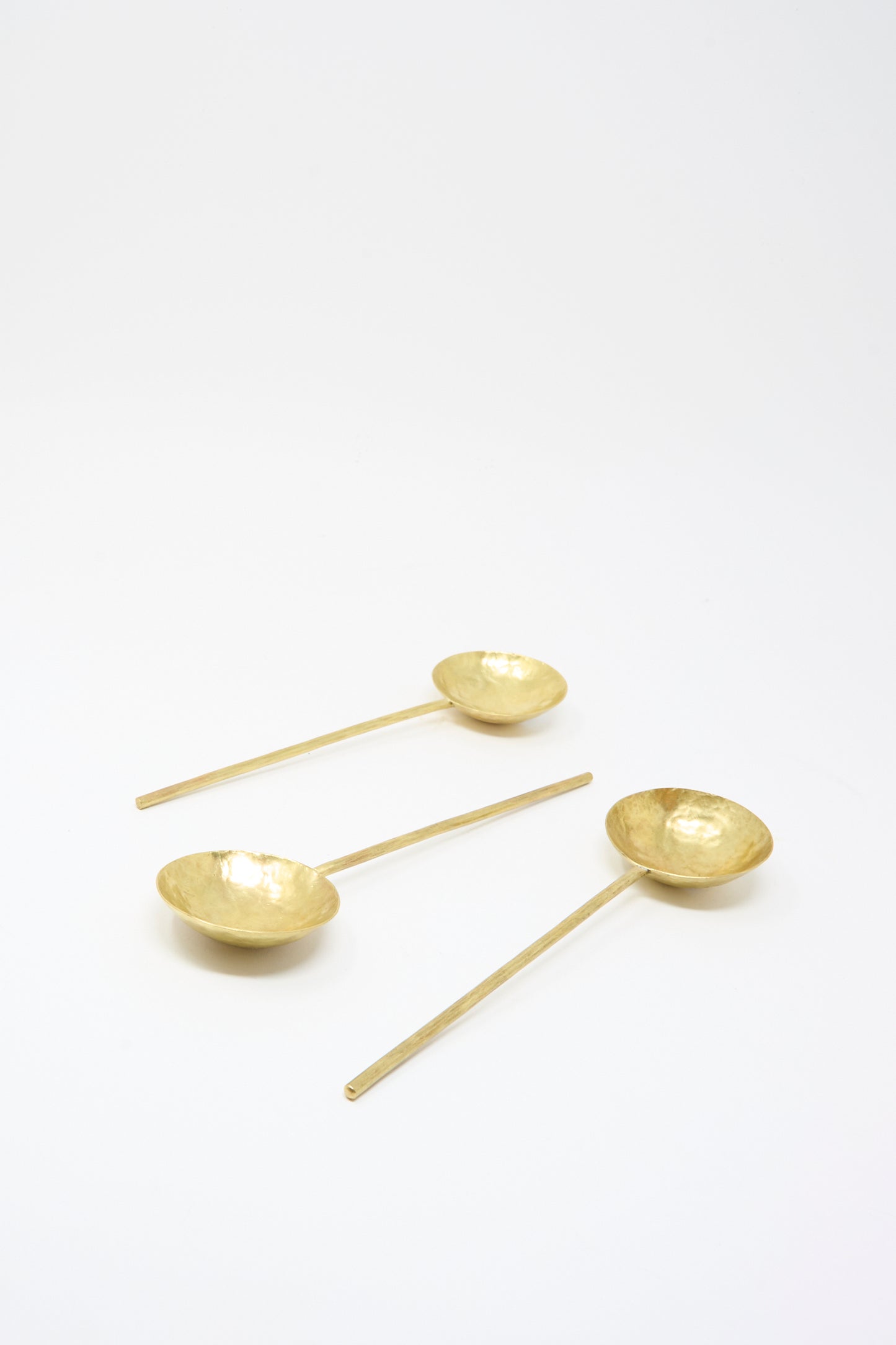 Three Totem Home Brass Serving Spoons, featuring gold-toned hammered solid brass and long handles, are arranged on a plain white background.