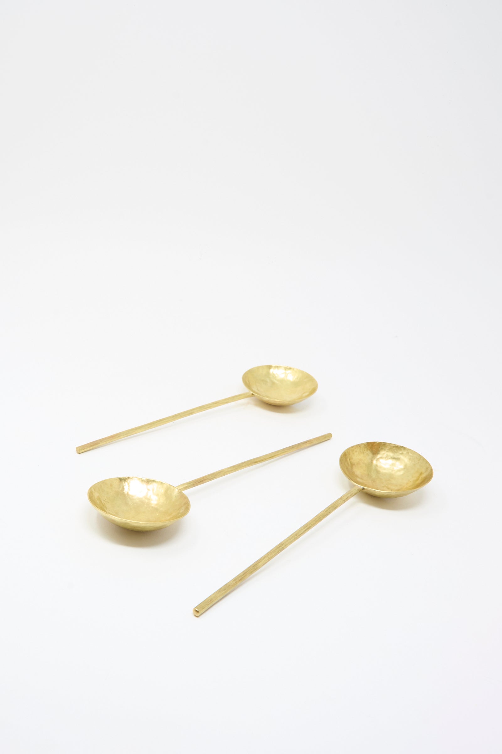 Three Totem Home Brass Serving Spoons, featuring gold-toned hammered solid brass and long handles, are arranged on a plain white background.