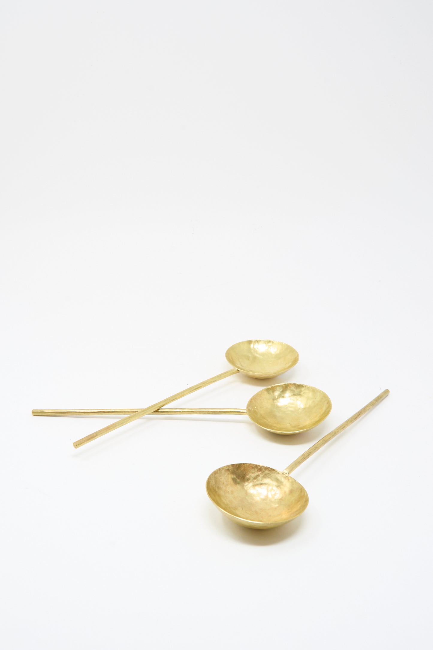 Three Totem Home Brass Serving Spoons, each gold-colored with hammered solid brass and long handles, are arranged diagonally on a plain white background.