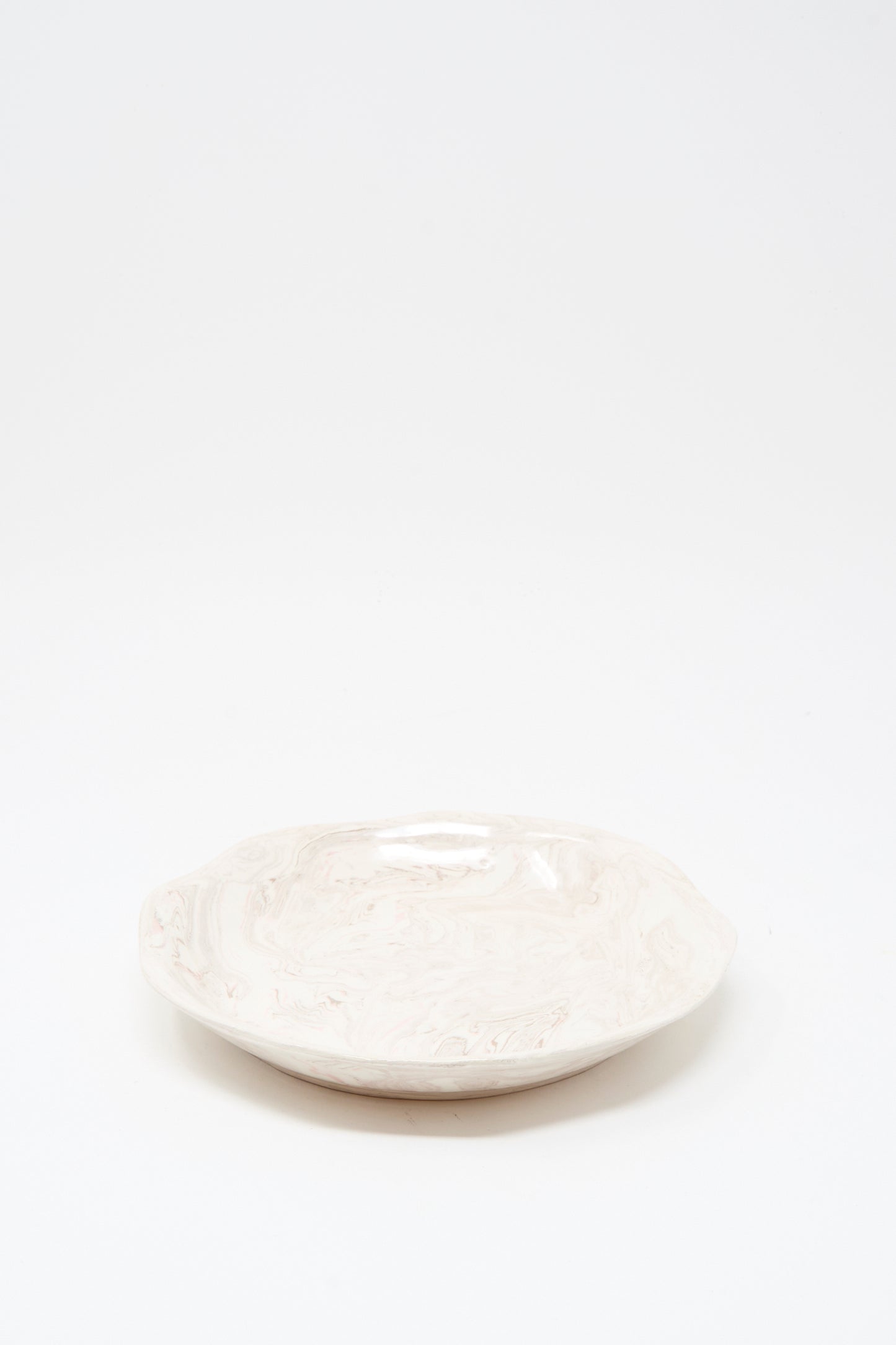 The Wild Rose Shallow Bowl by Totem Home is a handmade, off-white ceramic piece from Portugal, featuring a round shape, slightly wavy edge, and subtle pink marbled glaze, displayed against a plain white background.