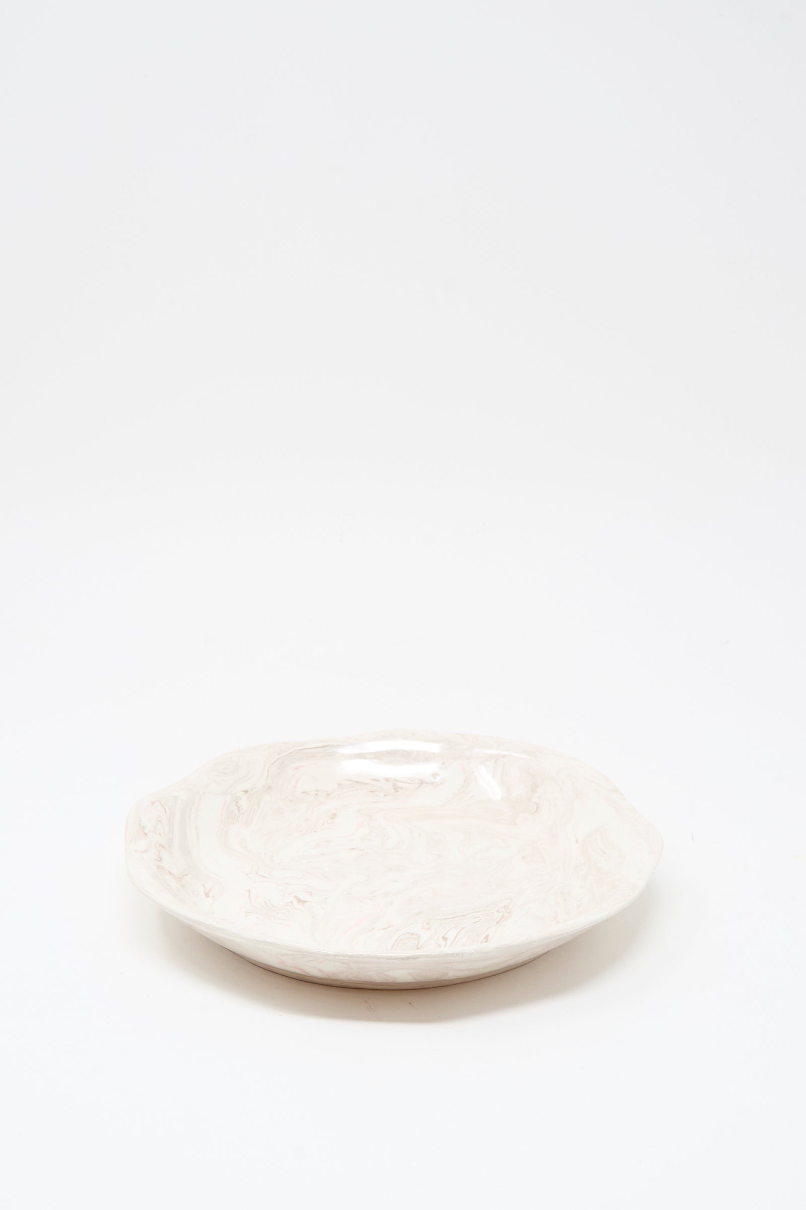 The Wild Rose Shallow Bowl by Totem Home is a handmade, off-white ceramic piece from Portugal, featuring a round shape, slightly wavy edge, and subtle pink marbled glaze, displayed against a plain white background.