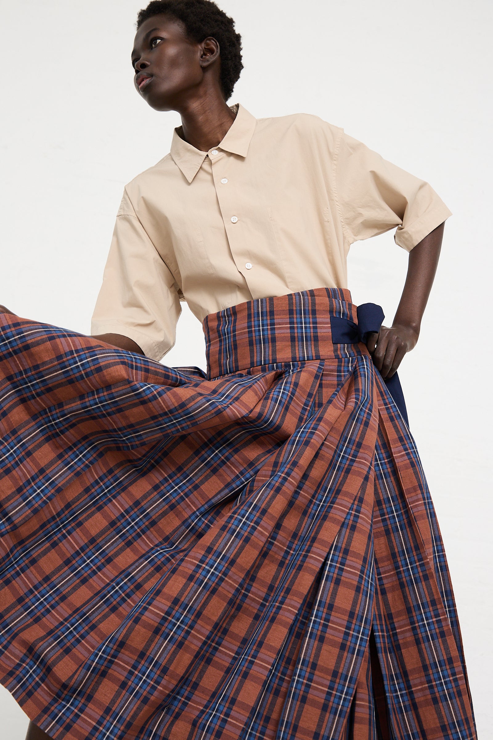 A person wearing a light beige short-sleeve button-up shirt and a Toujours Cotton Linen Madras Plaid Cloth Pleated Wrap Maxi Skirt in Brick posed against a plain white background.