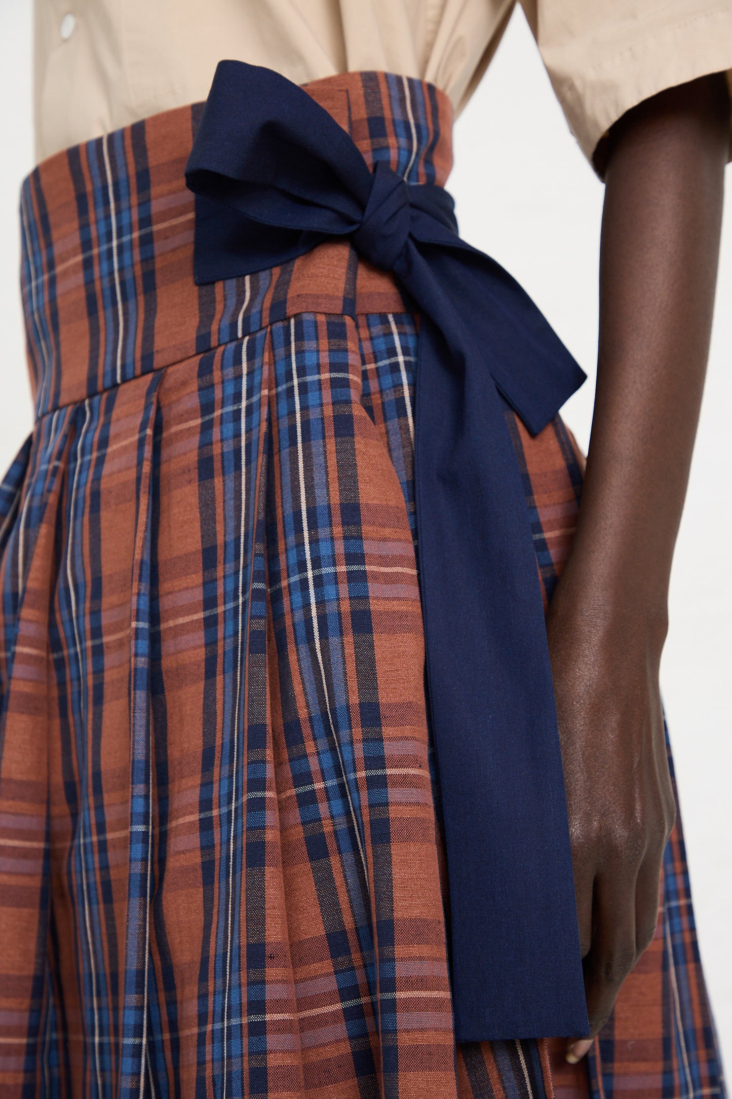Close-up of a person wearing the Toujours Cotton Linen Madras Plaid Cloth Pleated Wrap Maxi Skirt in Brick, featuring shades of brown and blue, pleats, and a wide waistband tied in a large dark blue bow. The person is wearing a beige top.