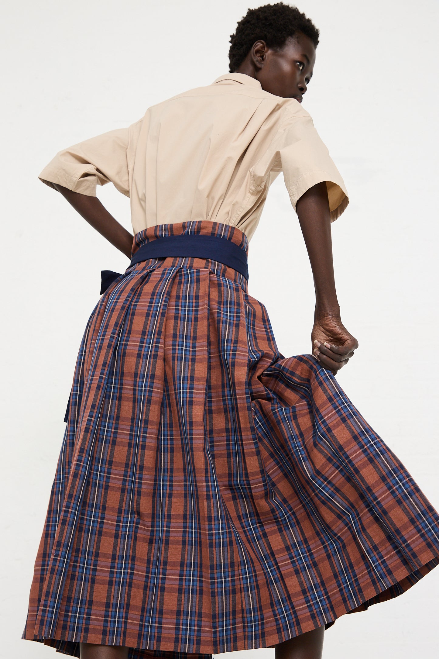 Person wearing a beige shirt and an a-line silhouette, Toujours Cotton Linen Madras Plaid Cloth Pleated Wrap Maxi Skirt in Brick, shown from the back, slightly lifting the skirt with their left hand.
