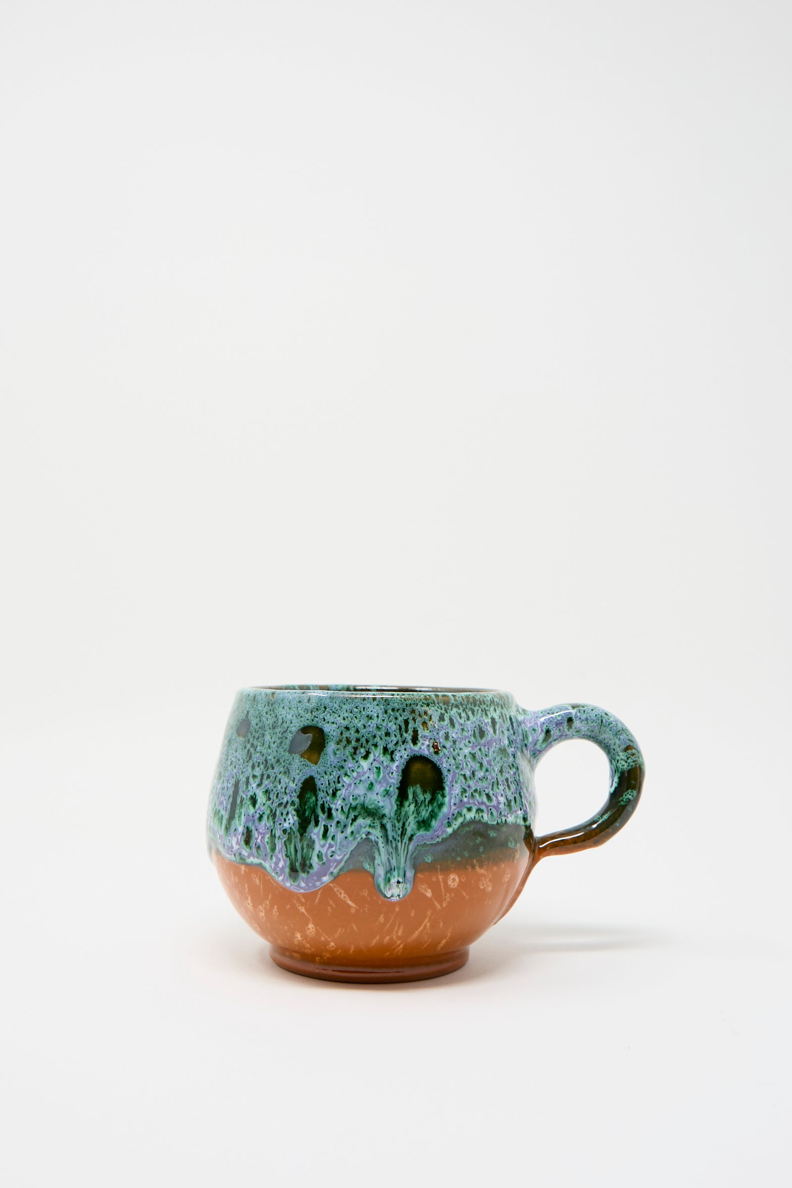 The Handmade Mug in Green and Orange from Travel Find showcases a round ceramic design with a green and purple glazed top and a brown base, against a white background. 