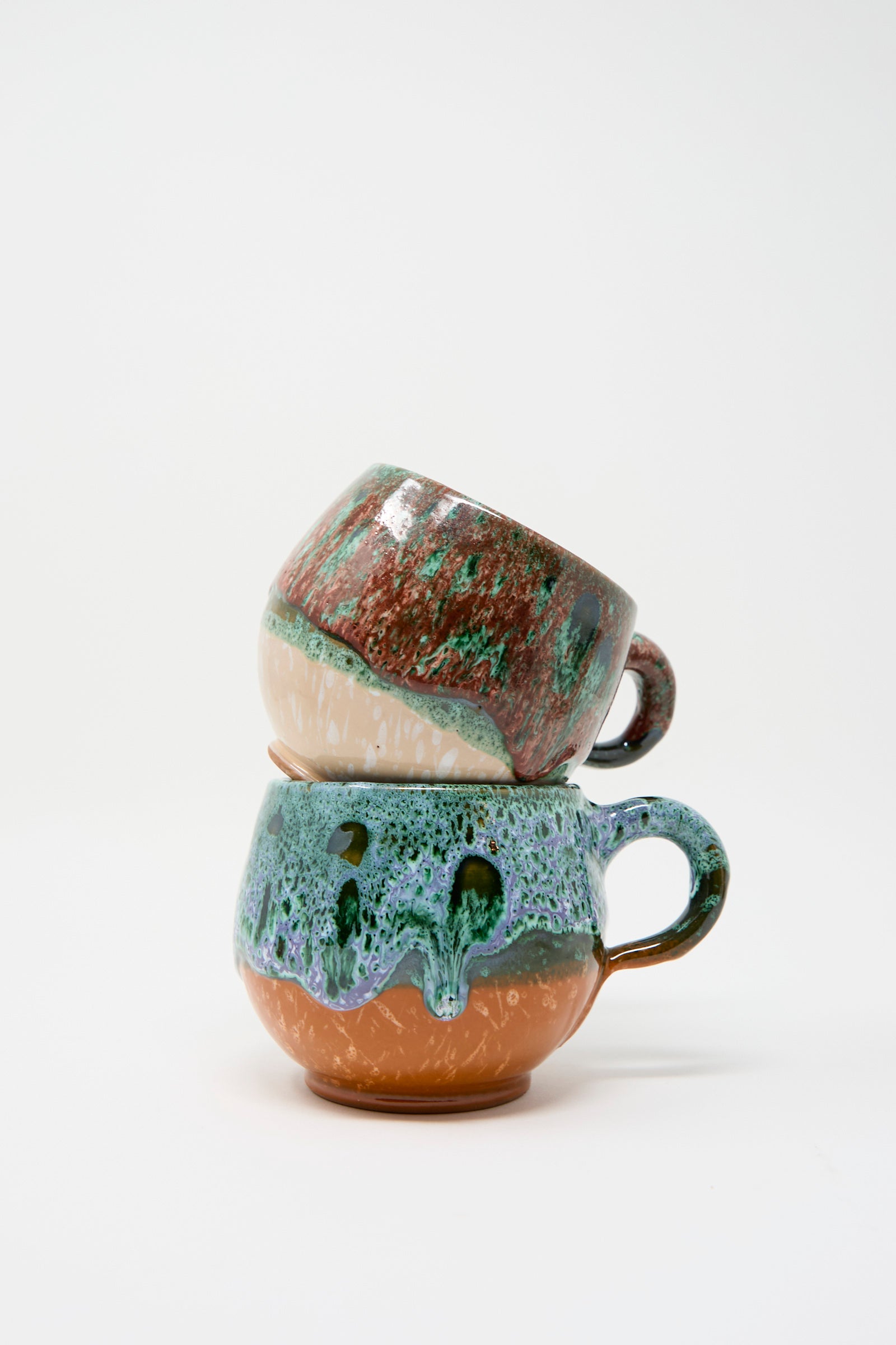 Two Travel Find Handmade Mugs in Green and Orange, showcasing a vibrant glaze with green, brown, and blue hues inspired by Moroccan aesthetics, are stacked on top of each other against a white background.