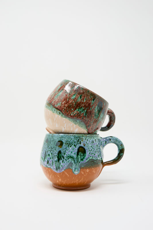 Two Travel Find Handmade Mugs in Green and Orange, showcasing a vibrant glaze with green, brown, and blue hues inspired by Moroccan aesthetics, are stacked on top of each other against a white background.