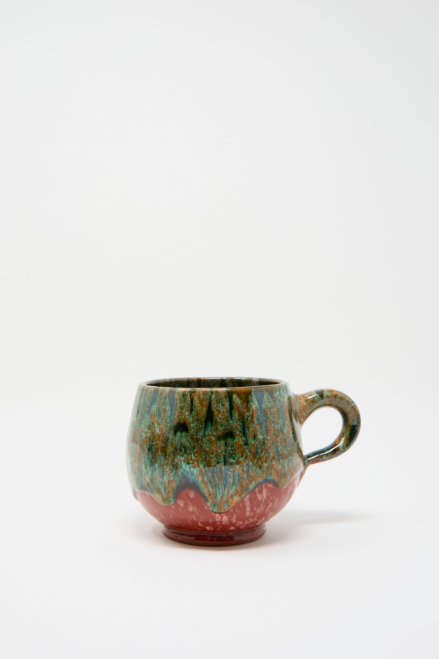 The "Handmade Mug in Green and Red" by Travel Find is a ceramic mug with a rounded shape, featuring a vibrant green, blue, and red glaze over a plain white background that reflects Moroccan clay artistry.