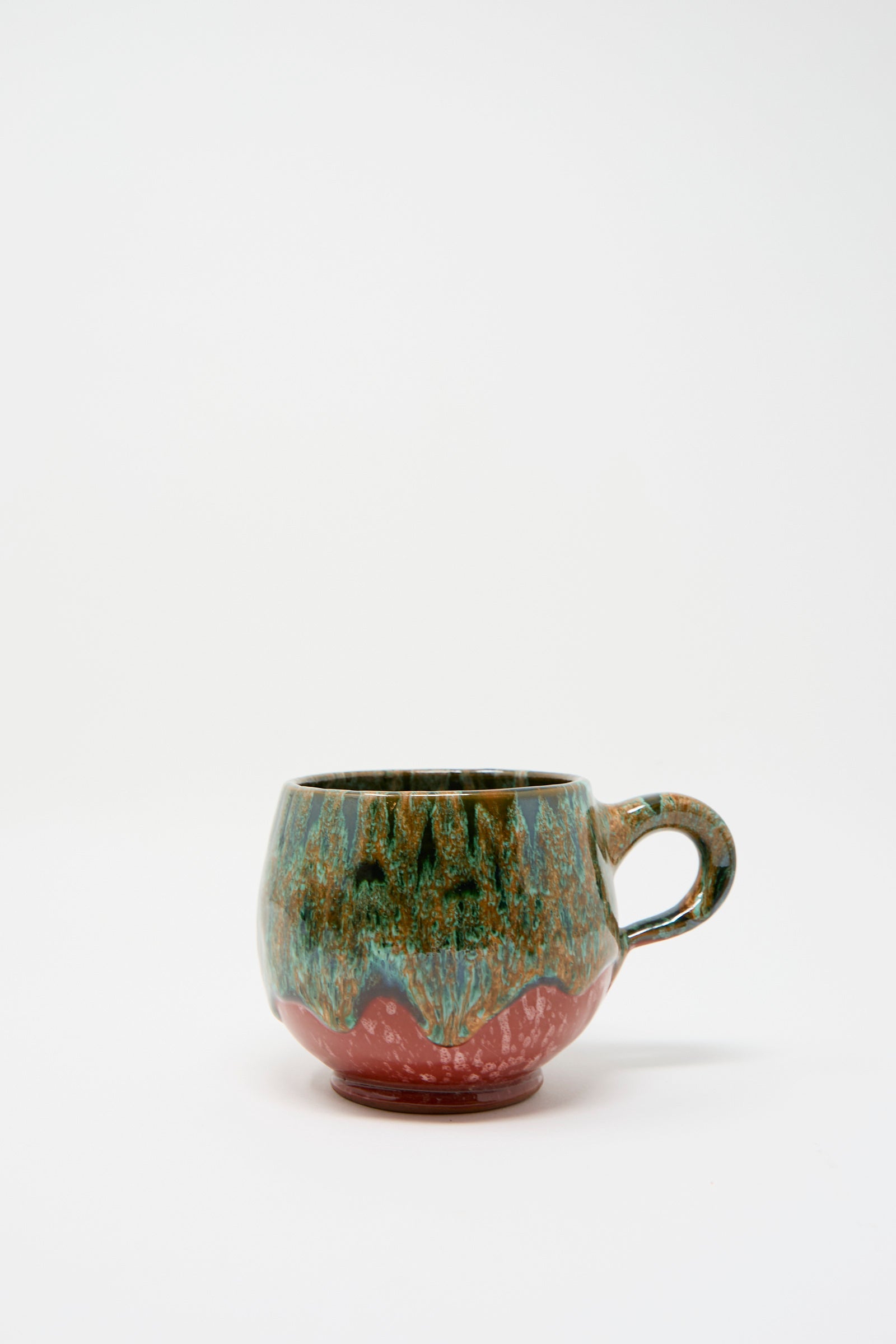 The "Handmade Mug in Green and Red" by Travel Find is a ceramic mug with a rounded shape, featuring a vibrant green, blue, and red glaze over a plain white background that reflects Moroccan clay artistry.