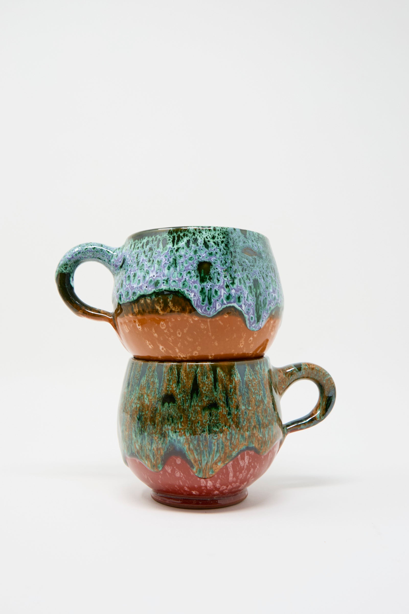 Two handmade ceramic mugs from Travel Find are stacked, each featuring a textured design. The top mug combines shades of blue and orange while the bottom mug blends green and brown. 