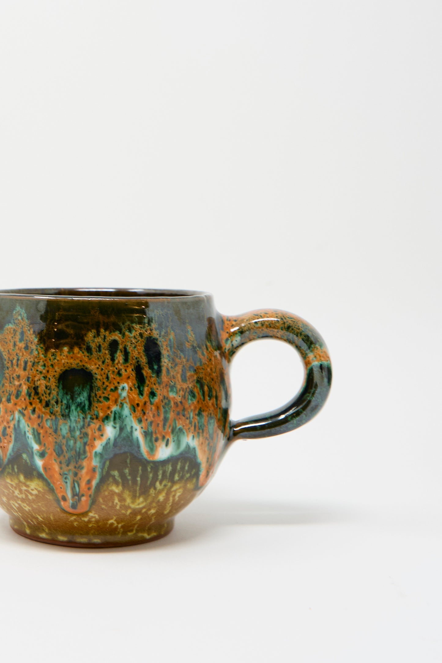 A close up of Travel Find's Handmade Mug in Green Orange Multi featuring a textured, multicolored glaze with shades of orange, blue, and green on a white background. 