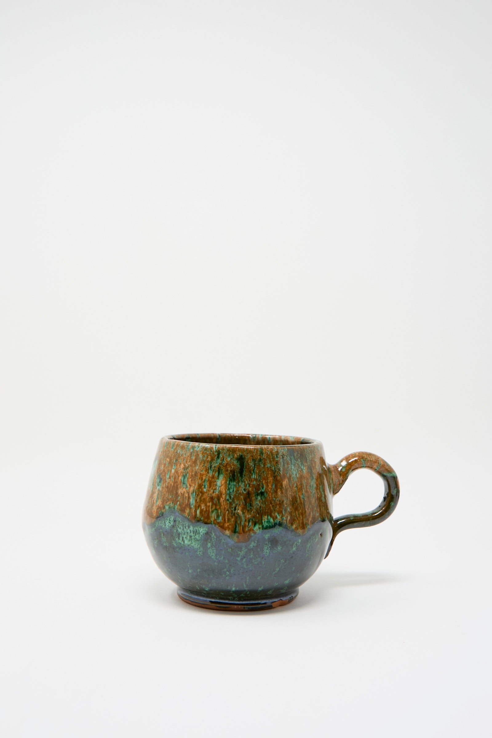 A rounded handmade ceramic mug from Travel Find, "Handmade Mug in Orange and Green," features a multi-color glaze of earthy browns and greens against a white background.