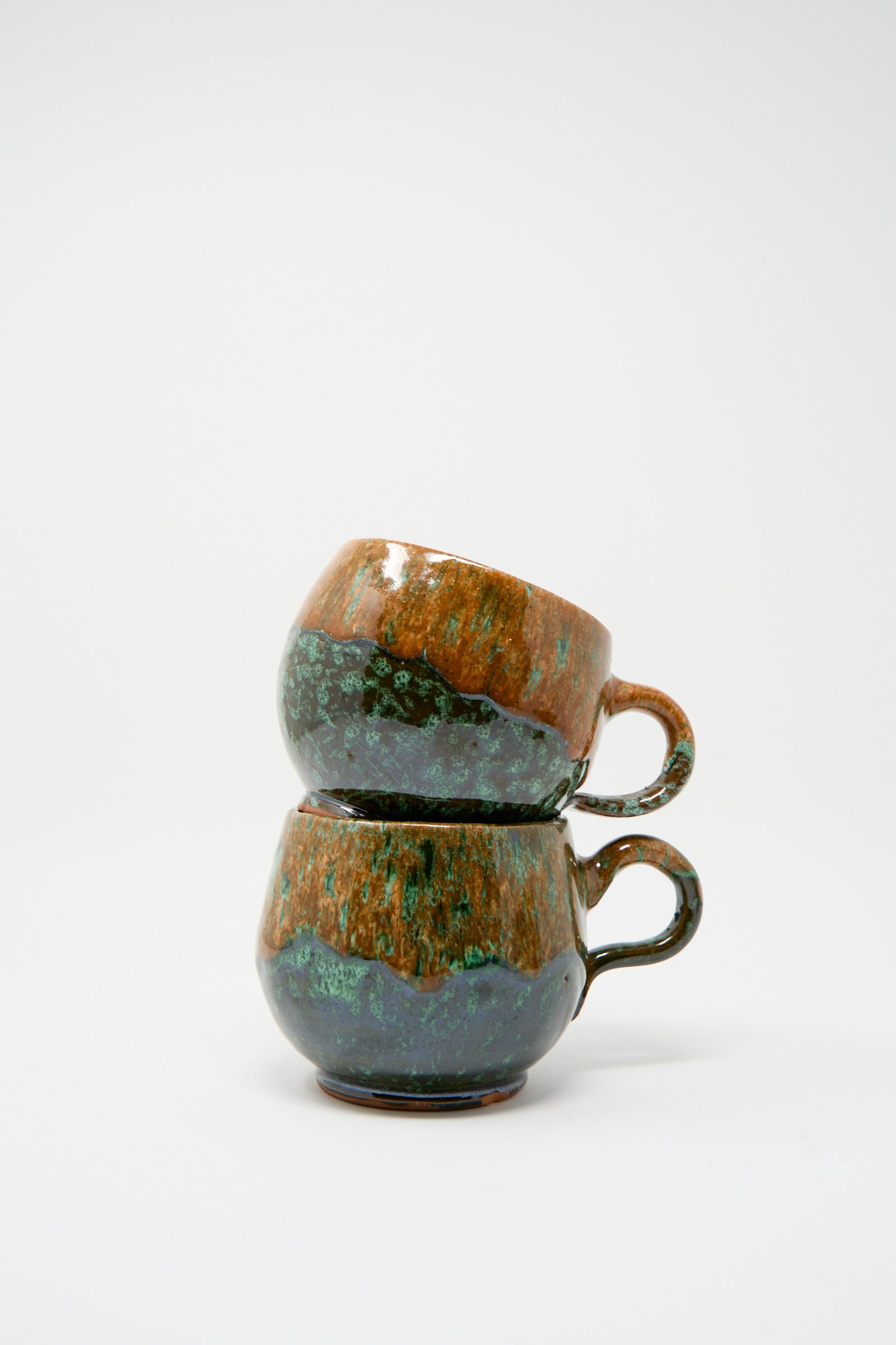 Two handmade ceramic mugs from Travel Find, each featuring a multi-color glaze of orange and green hues, are stacked against a plain white background.