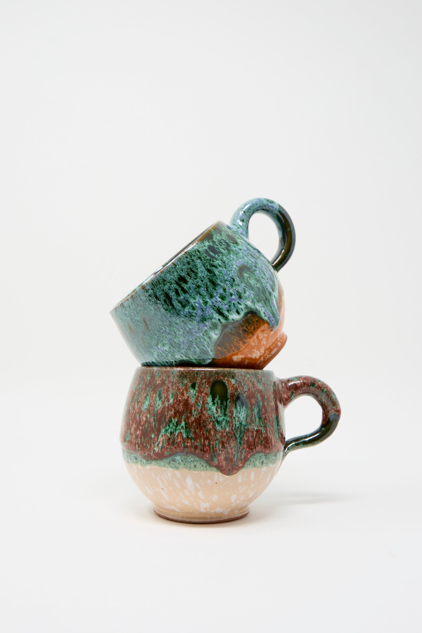 Two ceramic mugs from Travel Find, featuring a textured, multi-color glaze in green and red, are stacked against a plain white background. 