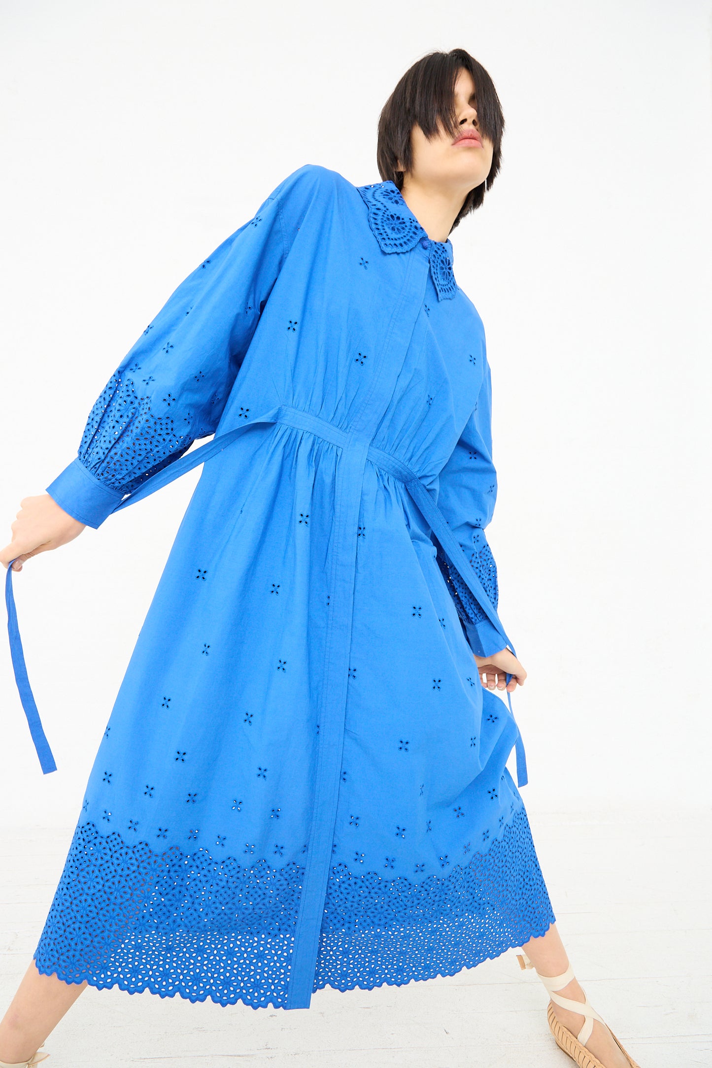 A woman in an Ulla Johnson Adette Dress in Cobalt with eyelet design details striking a dynamic pose against a white background.