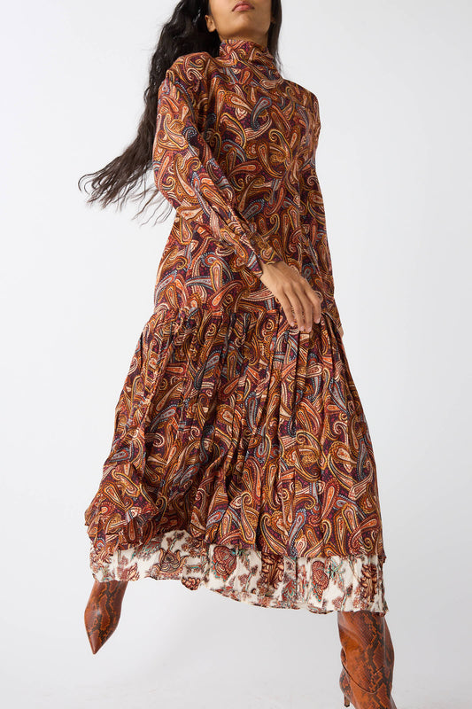 A person wearing the Adira Dress in Bergamot by Ulla Johnson, along with brown boots, stands against a plain white background. The paisley-patterned, long-sleeve turtleneck dress features a tiered, flowy skirt and a high neckline.