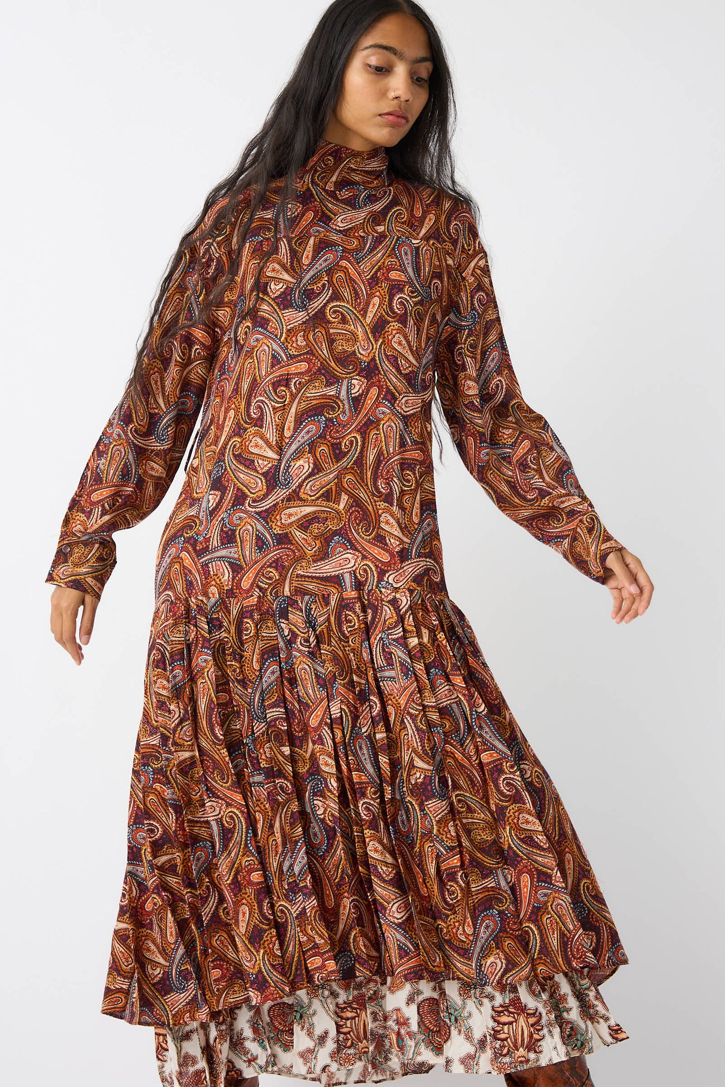 A person wearing the Adira Dress in Bergamot by Ulla Johnson, featuring a long-sleeved turtleneck design with a vibrant paisley pattern and layered hem, stands against a plain white background.