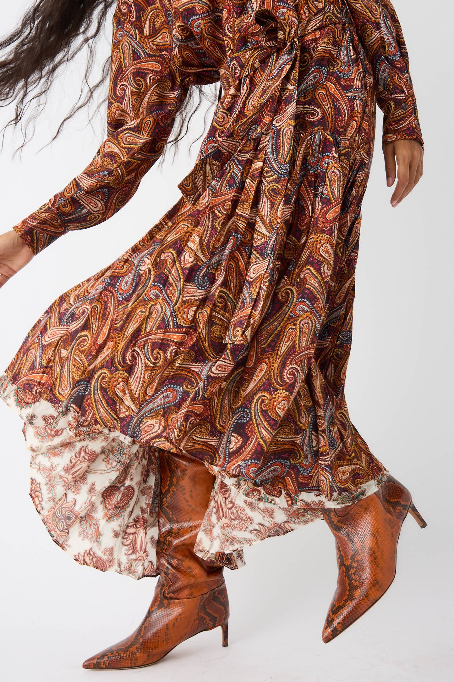 Person wearing the Ulla Johnson Adira Dress in Bergamot, featuring a flowing paisley-patterned turtleneck design, paired with knee-high brown boots in a dynamic pose. The hem of the dress reveals contrasting white fabric with red patterns.