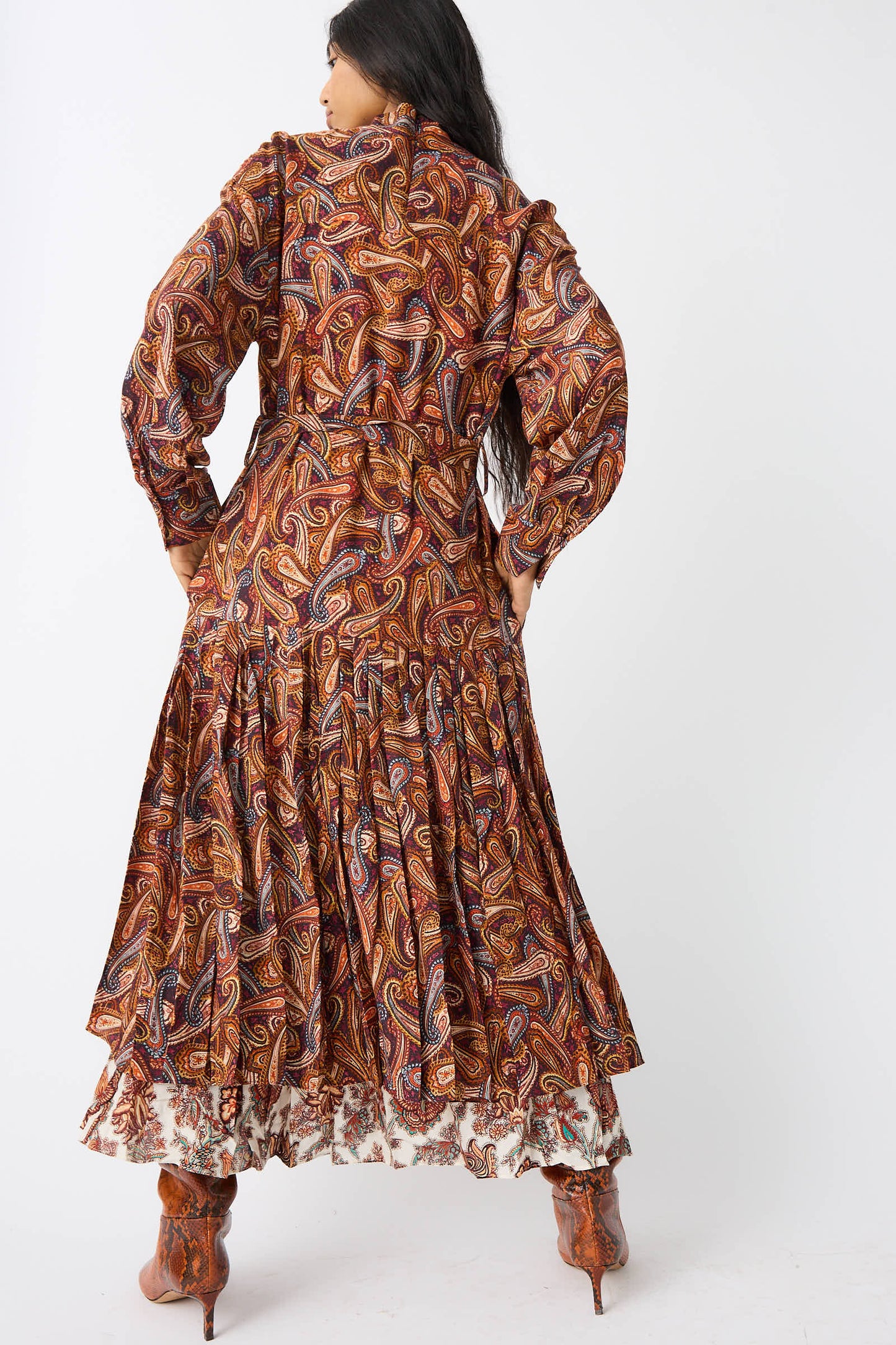 A person stands facing away, wearing the Ulla Johnson Adira Dress in Bergamot, which features a long-sleeve, multi-colored paisley pattern with a flowing, tiered skirt over a floral skirt and matching boots.