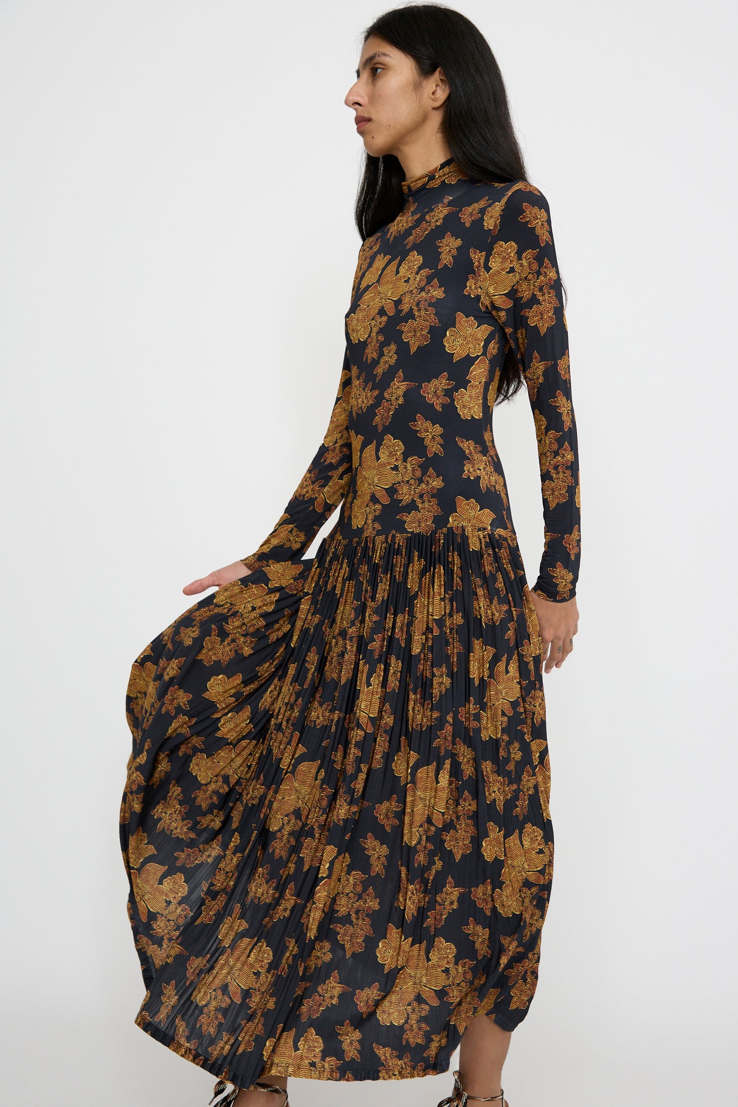 A person in the Ulla Johnson Amaya Dress in Onyx, featuring a floral pattern, long sleeves, a pleated skirt, and a subtle turtleneck, stands against a plain background.