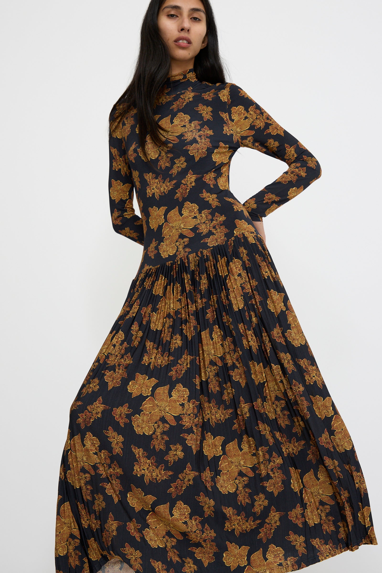 A person wearing the Amaya Dress in Onyx by Ulla Johnson, featuring long sleeves and gold floral patterns, stands against a plain background.