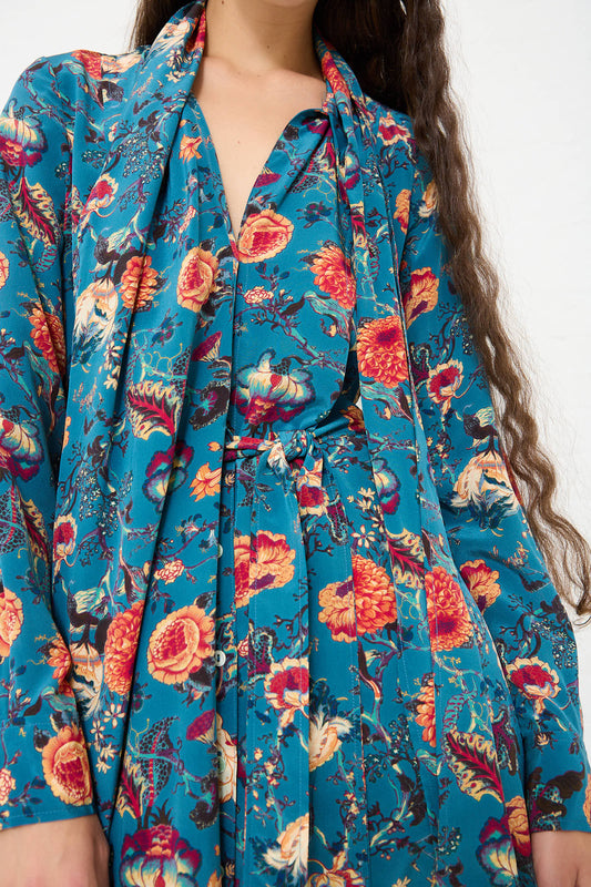 A person wearing the Ulla Johnson Anisa Maxi Dress in Bluette with a tie at the collar and waist. The long-sleeved silk maxi dress features vibrant orange and yellow botanical print flowers. The person has long, wavy hair. 