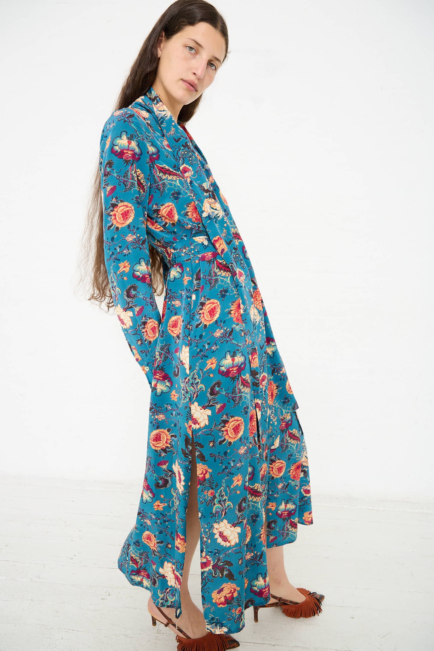 A person with long hair stands in an Ulla Johnson Anisa Dress in Bluette featuring a teal botanical print and brown moccasins against a plain white background.