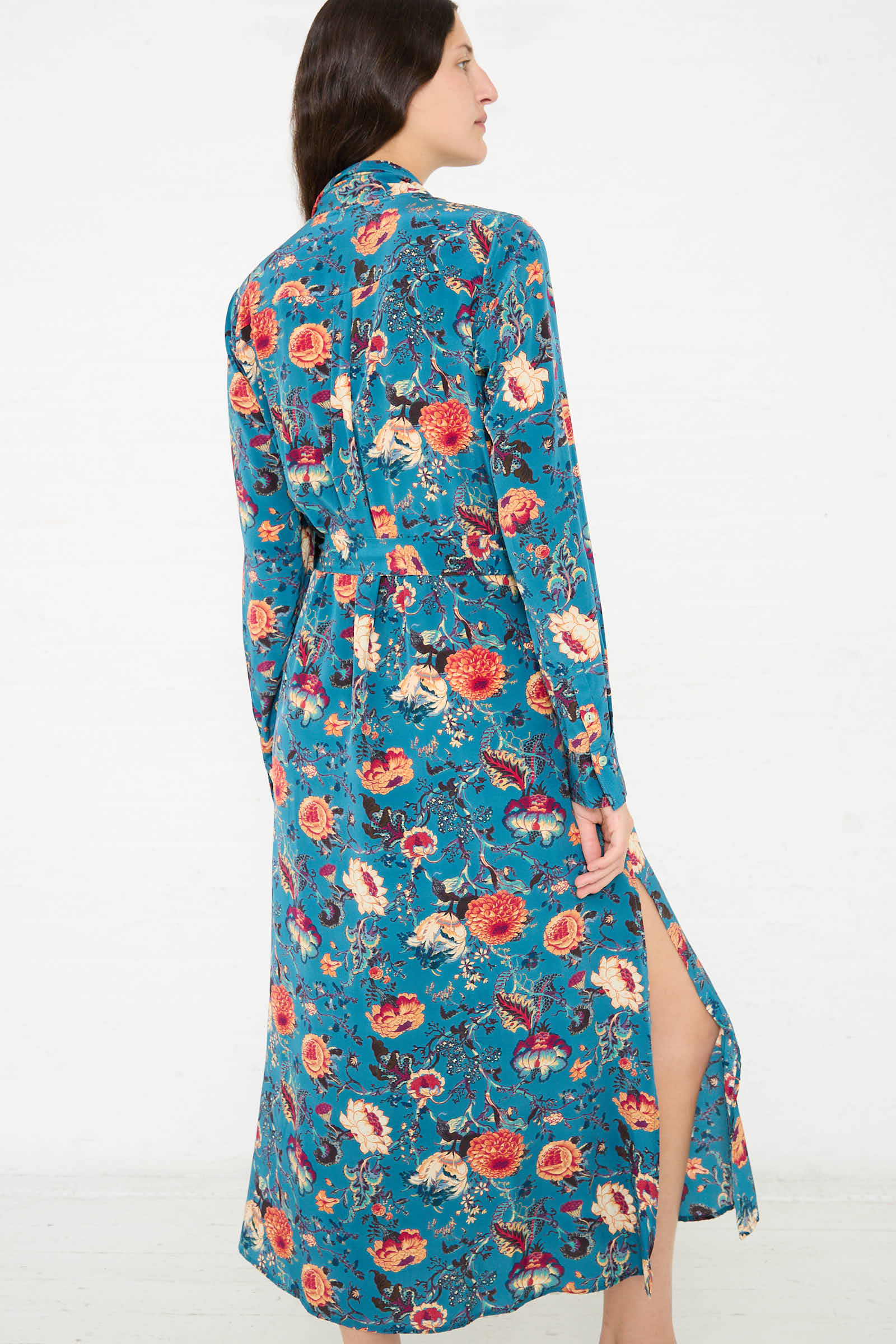 A woman stands facing sideways in the Ulla Johnson Anisa Dress in Bluette, a vintage-inspired blue floral silk maxi dress with a front slit, against a plain white background.