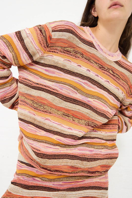 Person wearing a multicolored, striped, long-sleeved Ulla Johnson Ansel Pullover in Sunset. The relaxed fit sweater features various shades of orange, pink, and brown.