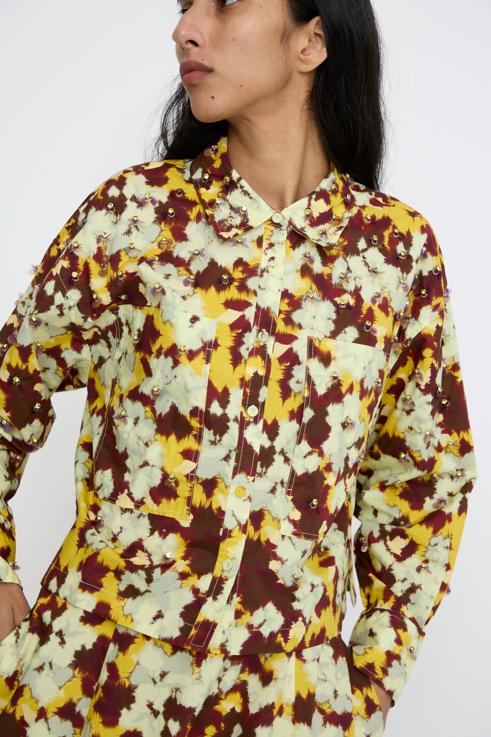 A model dressed in the Ulla Johnson Aria Blouse in Acacia, featuring a yellow, brown, and white tie-dye pattern, glances to the side against a plain background.