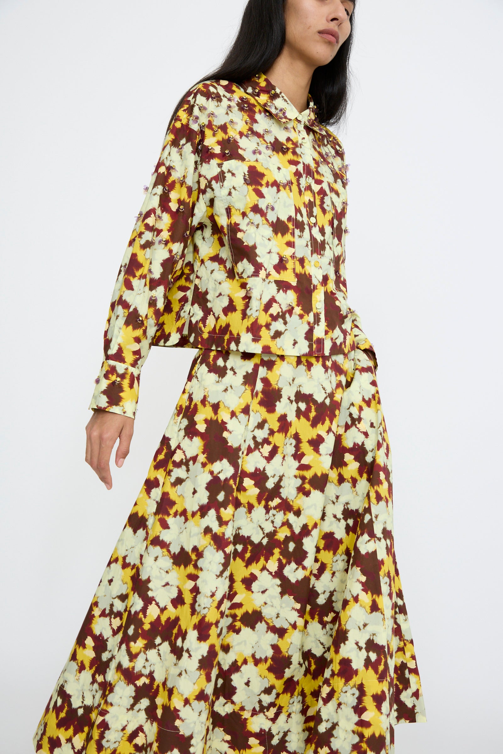 A person wearing a floral-patterned ensemble by Ulla Johnson, featuring a long-sleeve top and the Vianne Skirt in Acacia, showcasing shades of yellow, white, and maroon against a white background