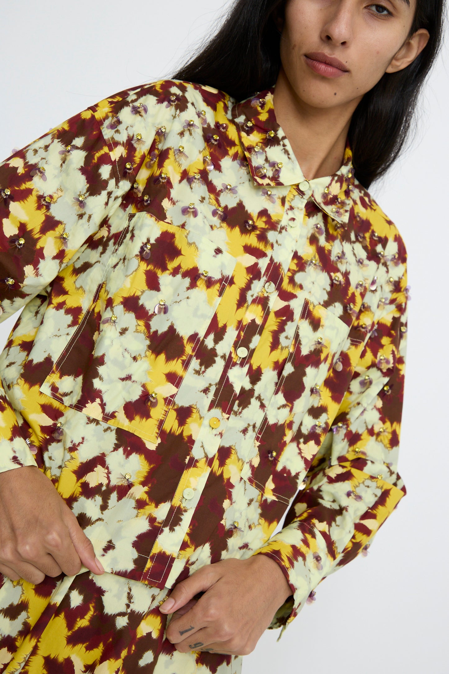 A person wearing the Ulla Johnson Aria Blouse in Acacia, featuring a floral pattern in shades of yellow, brown, and white with bead embellishments, looks at the camera while adjusting the hem.