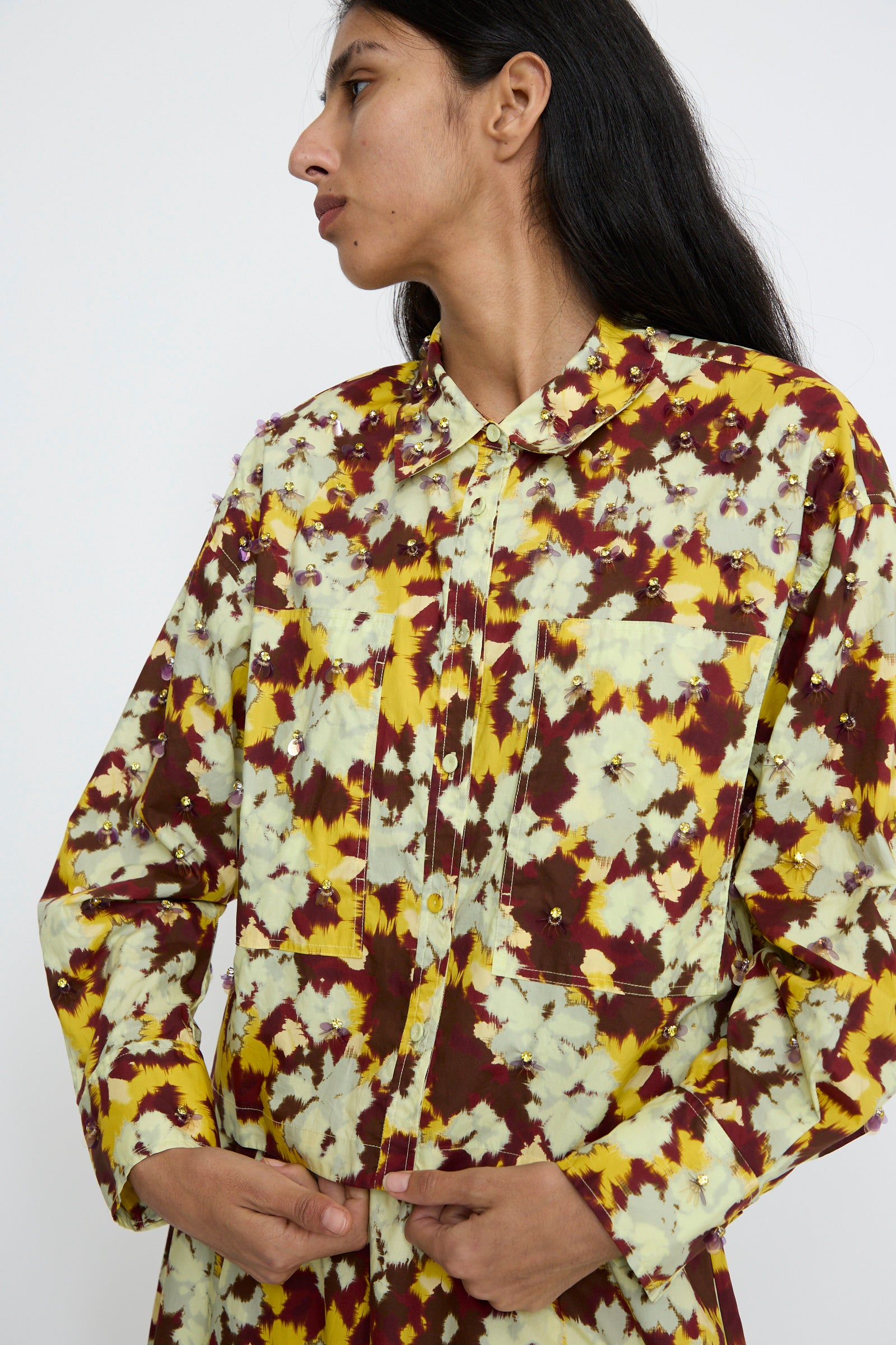 A person is wearing the Ulla Johnson Aria Blouse in Acacia, featuring a regular fit with long sleeves and an ikat dyed cotton pattern of yellow, maroon, and white florals, gazing to the side.